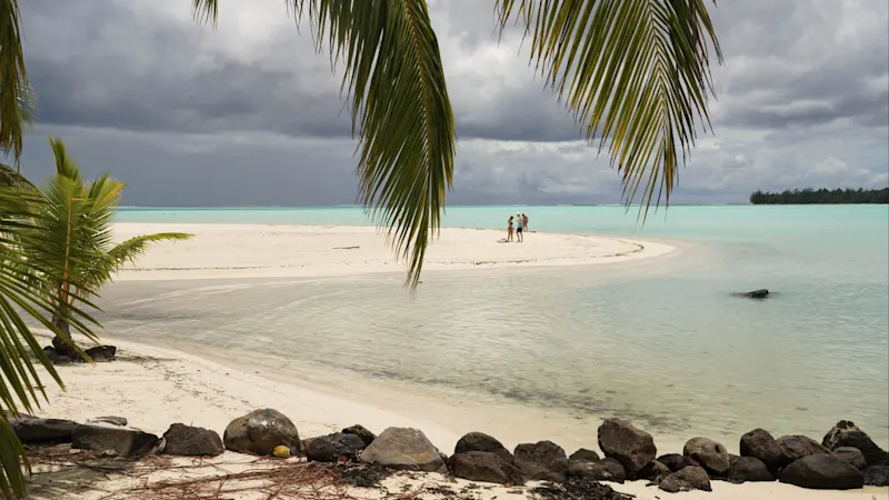 Tropischer Sandstrand mit türkisfarbenem Wasser, Palmenblättern und dunklen Wolken. Menschen stehen auf einer Sandbank.