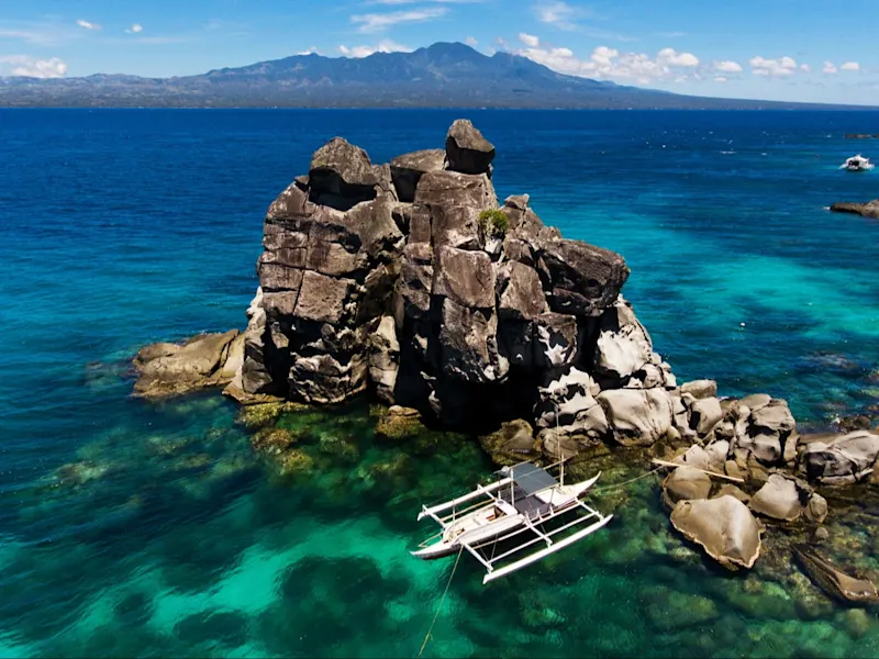 Apo Island, Philippinen. Felsformation im türkisfarbenen Meer mit Boot, im Hintergrund Berge, Apo Island, Philippinen.