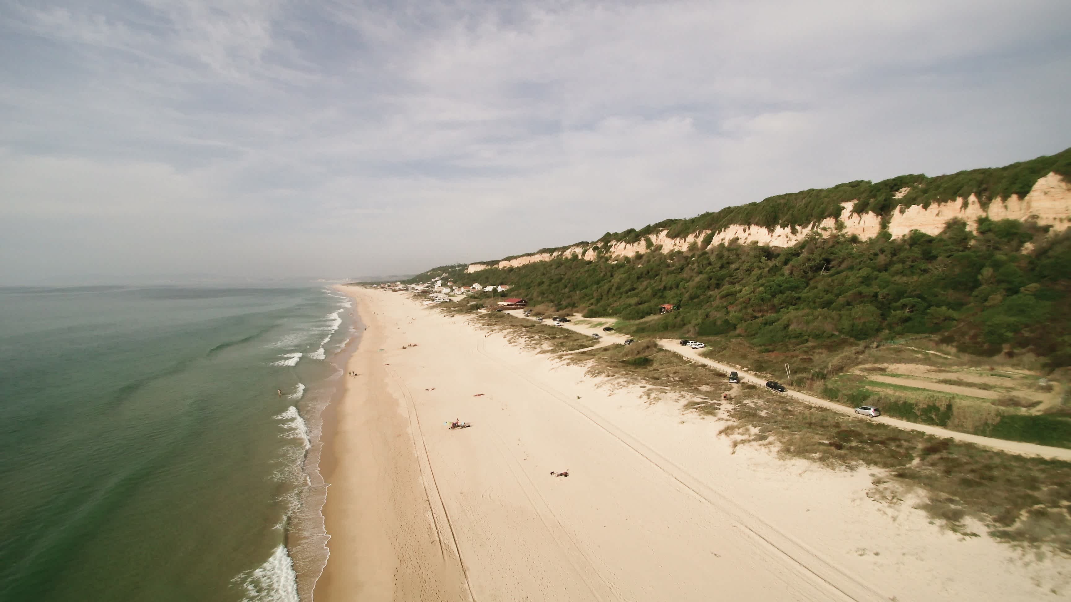 Der lange Sandstrand von Praia da Fonte da Telha, Portugal mit grünen Dünen bei bewölktem Wetter.