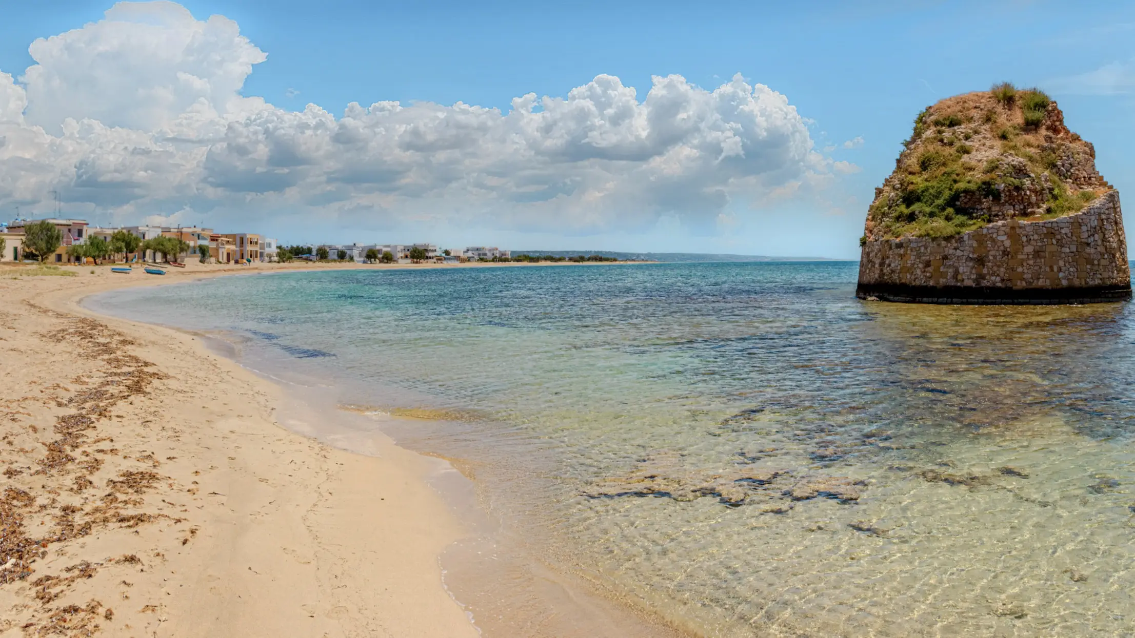 Mediterraner Sandstrand mit türkisblauem Wasser, Felsformation im Meer und Küstenort unter weißen Wolken am blauen Himmel.