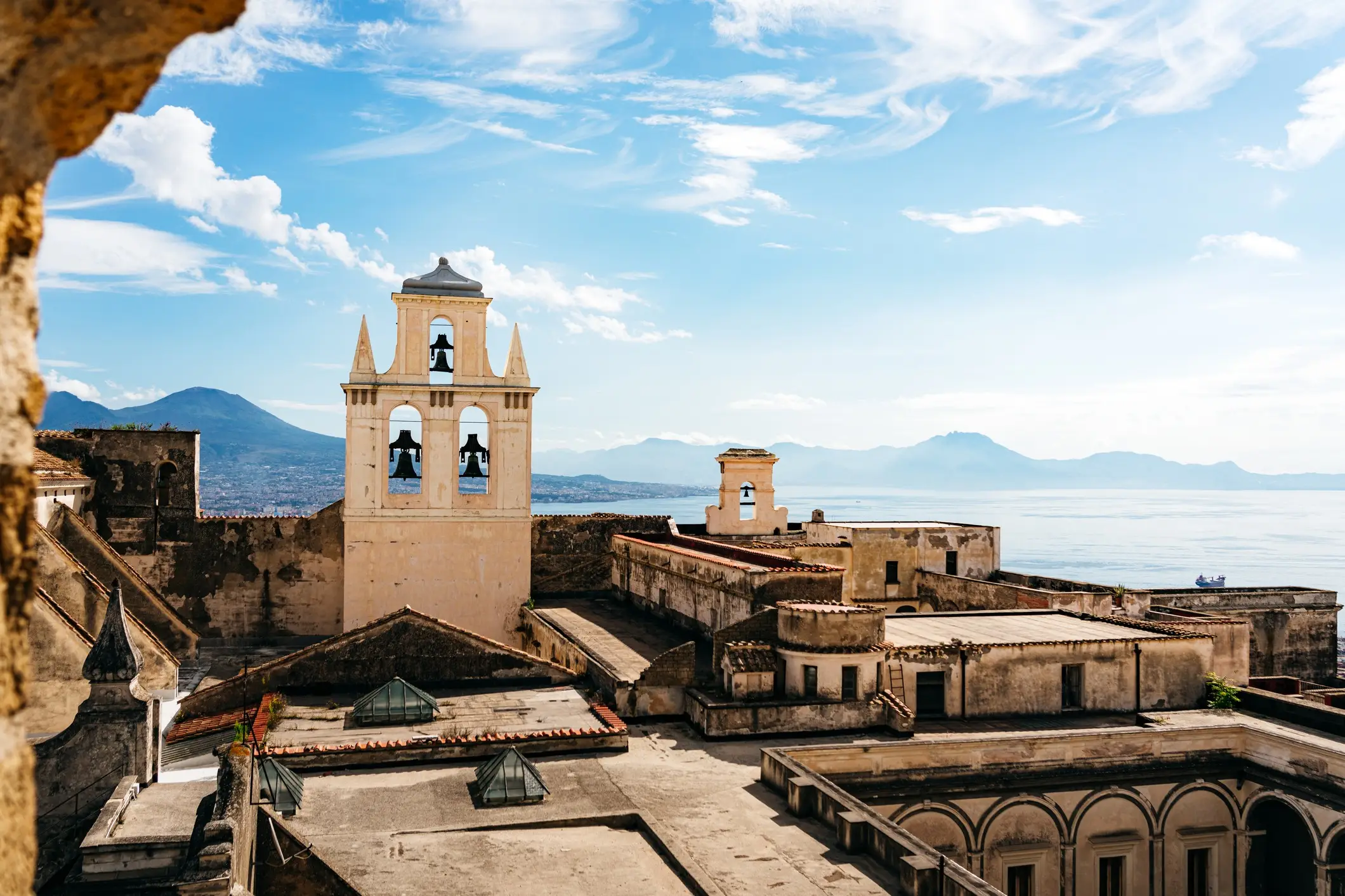 Vue panoramique d'un monastère méditerranéen avec clocher, surplombant la baie et les montagnes sous un ciel bleu.