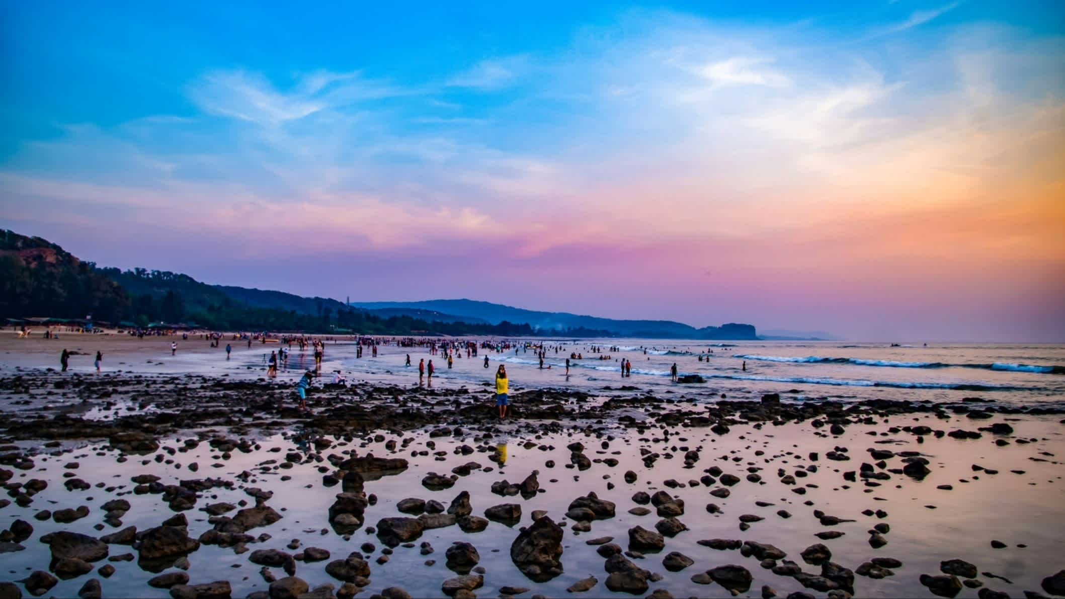 Bunter Sonnenuntergang am Kashids Strand, Maharashtra, Indien mit Menschen im Wasser bei Ebbe und freigelegten Felsen.