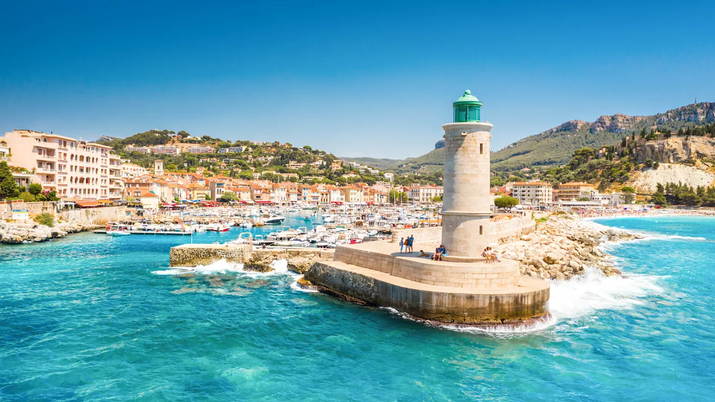 Stone lighthouse with turquoise dome on harbor pier, overlooking Mediterranean marina and coastal village.