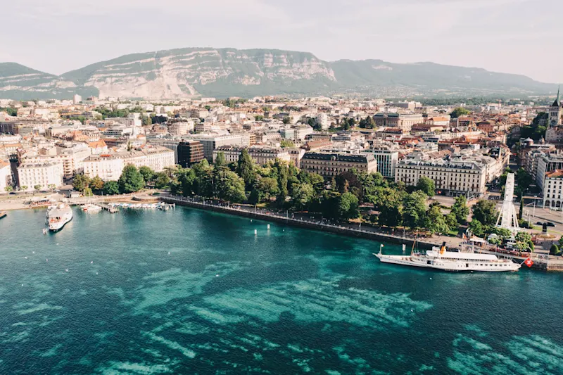 Port, city center, Geneva Aerial view of Geneva with turquoise Lake Geneva waters, boats at harbor, city buildings, and mountains in background.