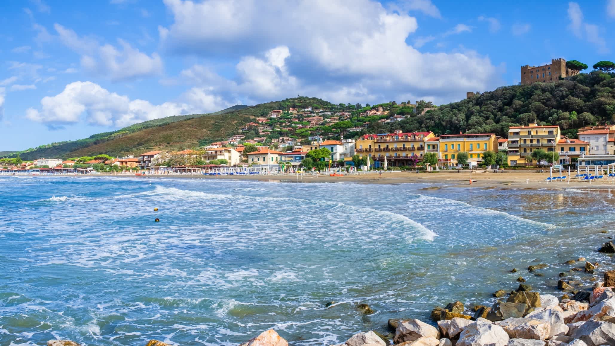 Meereslandschaft in Castiglione della Pescaia, Toskana, Italien mit Häusern am Rand des Strandes und Bergen im Hintergrund.

