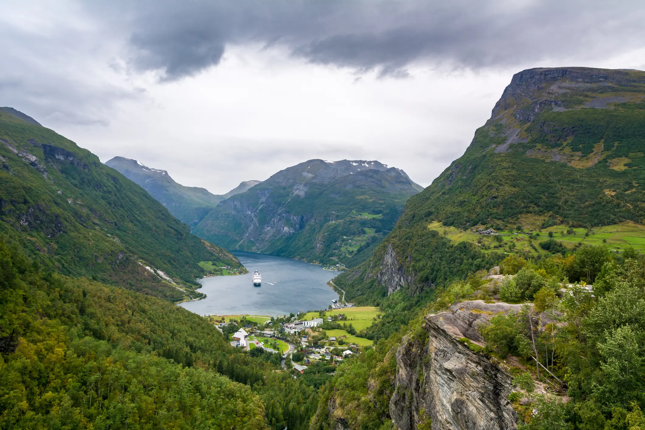 Ein malerischer Blick auf einen Fjord, der von steilen, grünen Bergen umgeben ist, mit einer kleinen Stadt und einem Kreuzfahrtschiff im Wasser darunter, unter einem bewölkten Himmel.