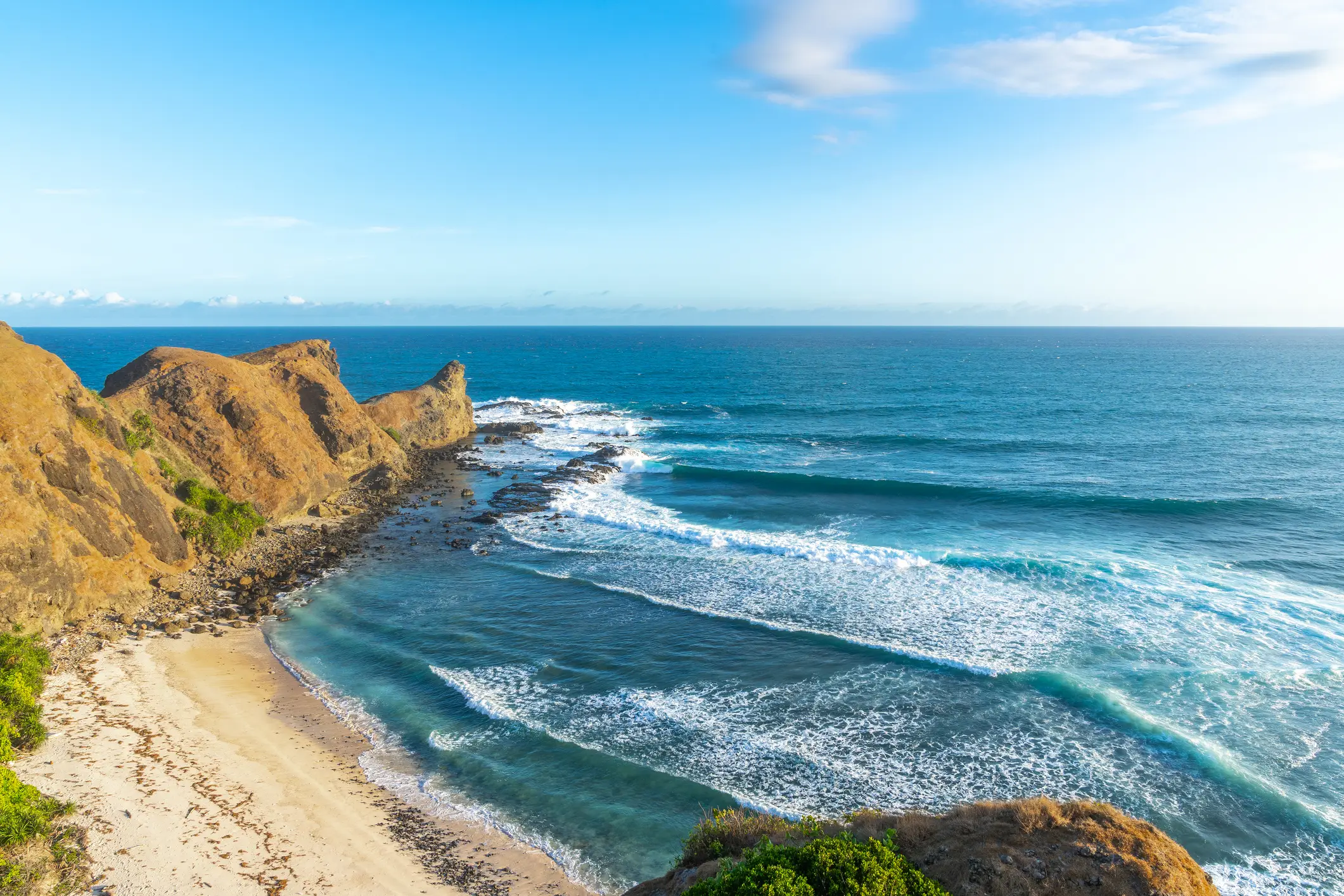 Felsige Küstenlandschaft mit türkisblauem Meer, sandigem Strand und schäumenden Wellen unter klarem Himmel.