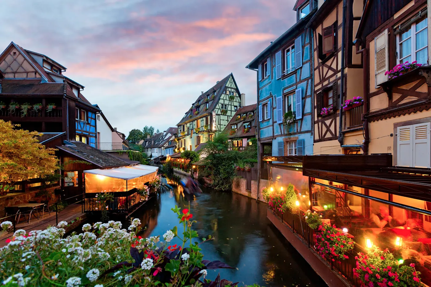 Colorful half-timbered buildings along a canal in Colmar, France at twilight with flowers and illuminated restaurants.