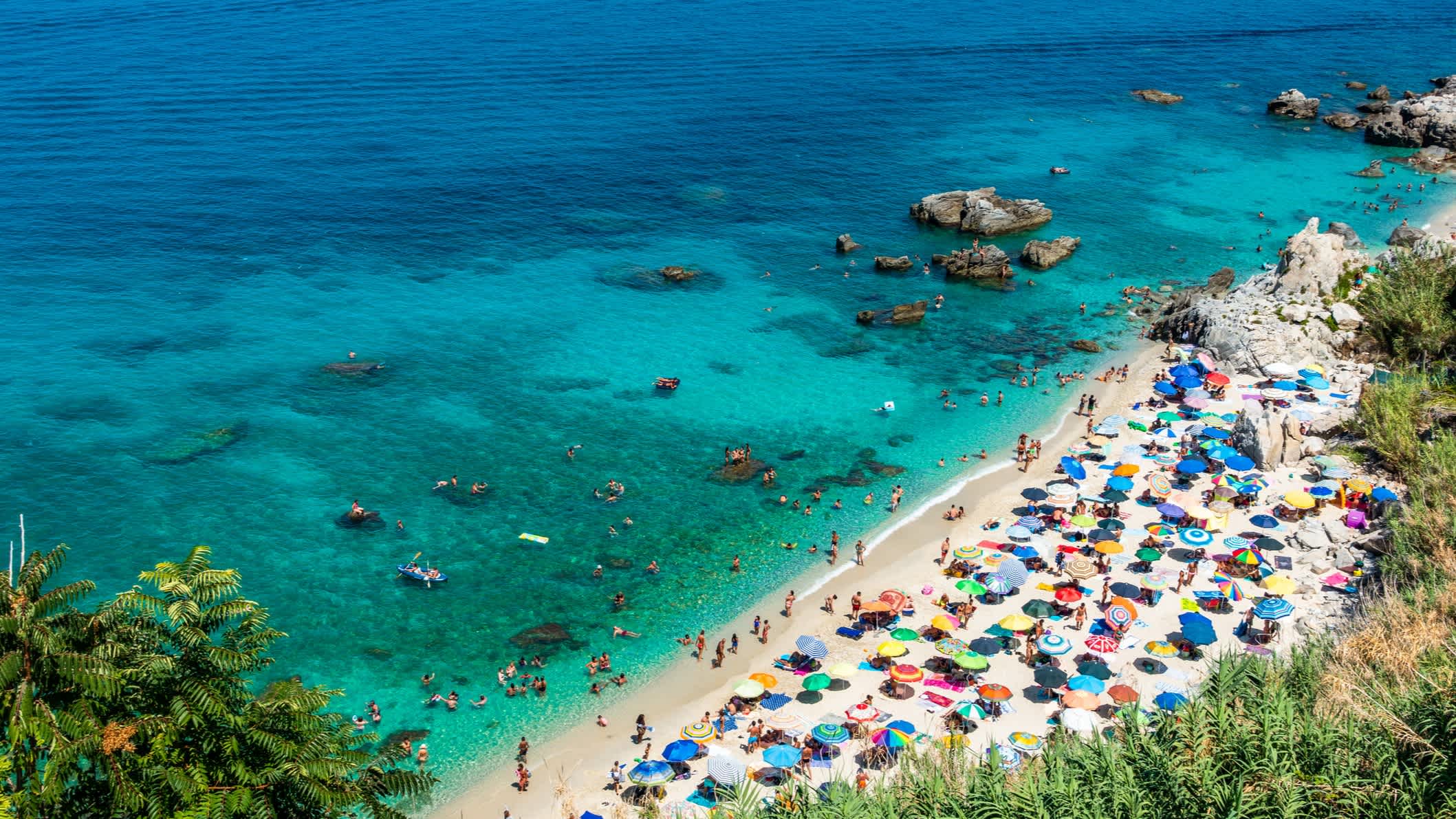 Blick auf den Strand von Parghelia, Kalabrien, mit bunten Sonnenschirmen, eingerahmt von üppiger Vegetation und kristallklarem Wasser.
