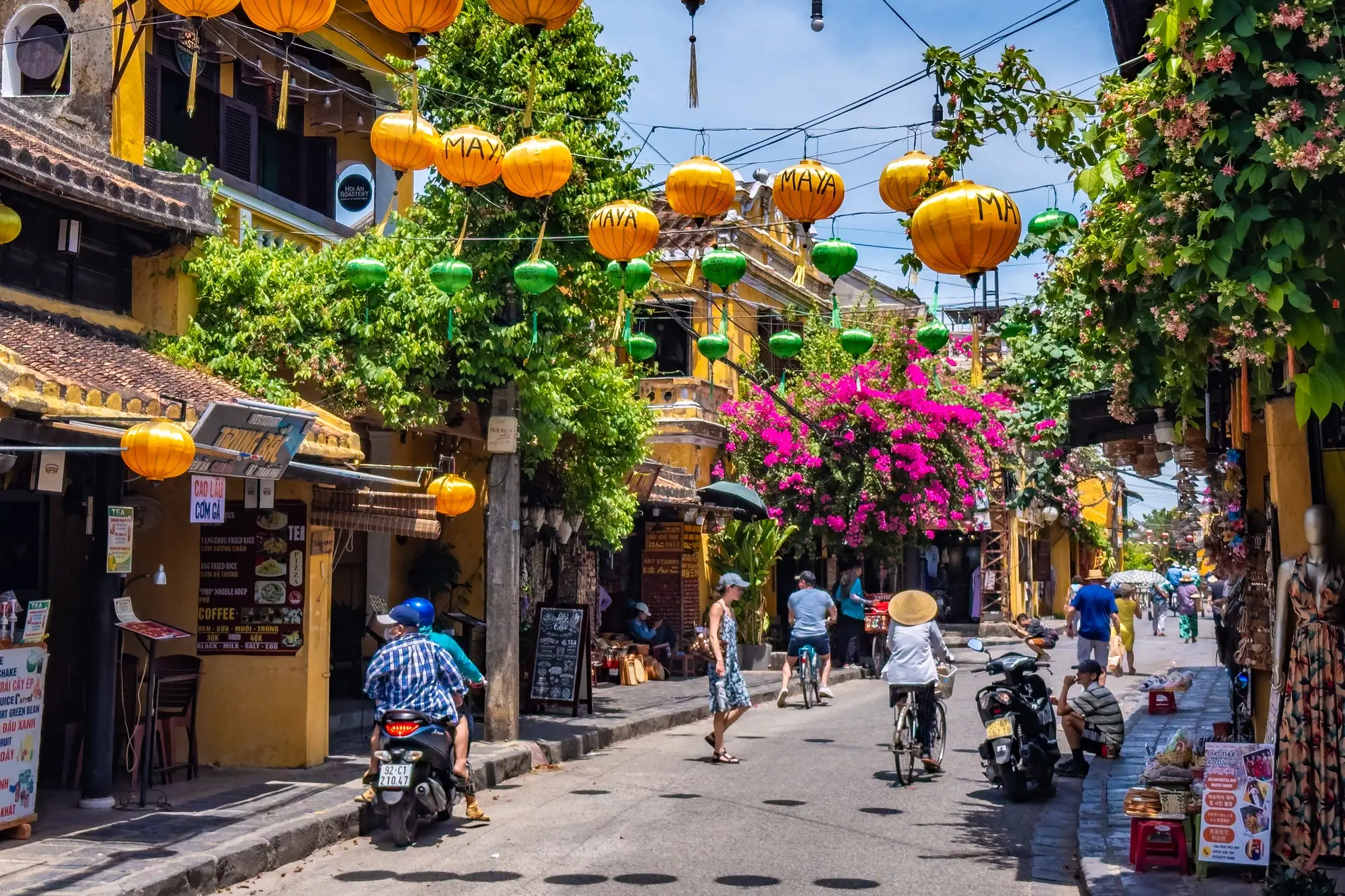 Bunte Straße in Hoi An mit gelben und grünen Laternen, blühenden Pflanzen und Touristen auf Fahrrädern und Motorrollern.