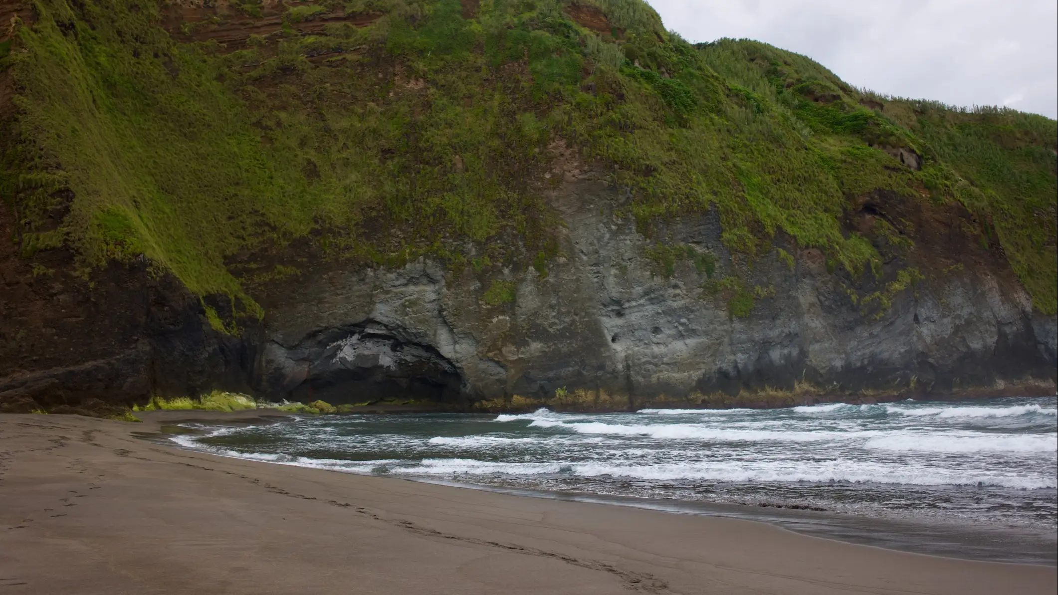 Grünes Gras an der Klippe von Praia do Areal de Santa Bárbara, São Miguel, Azoren, Portugal mit Blick auf eine kleine Insel im Wasser.