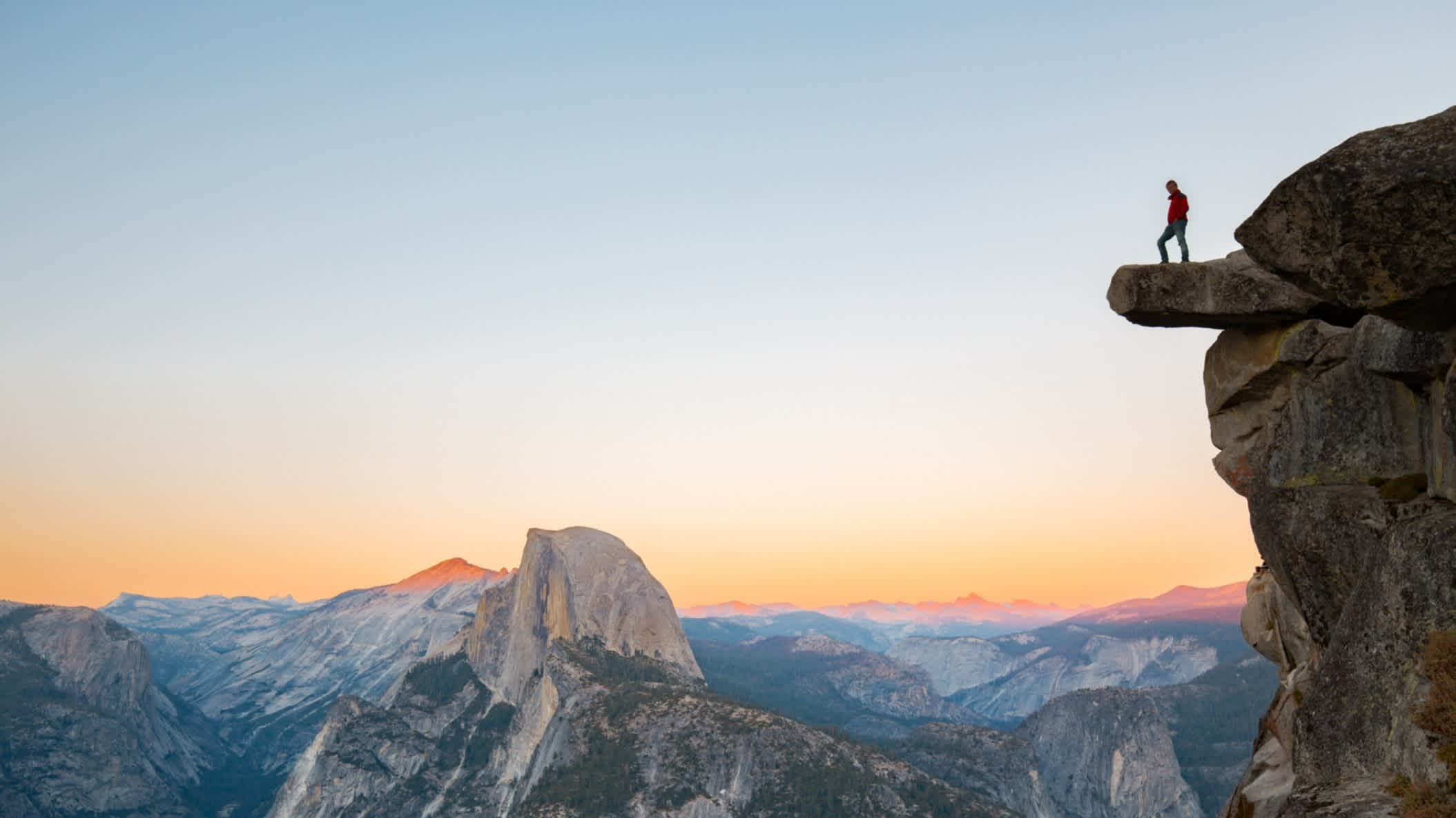 Mann auf Klippe überblickt Gebirge bei Sonnenaufgang.