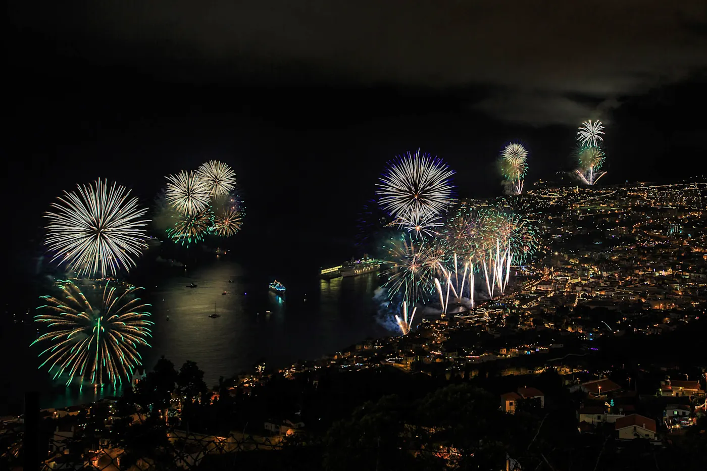 Colorful fireworks display over a coastal city at night, illuminating the bay and surrounding hillside neighborhoods.