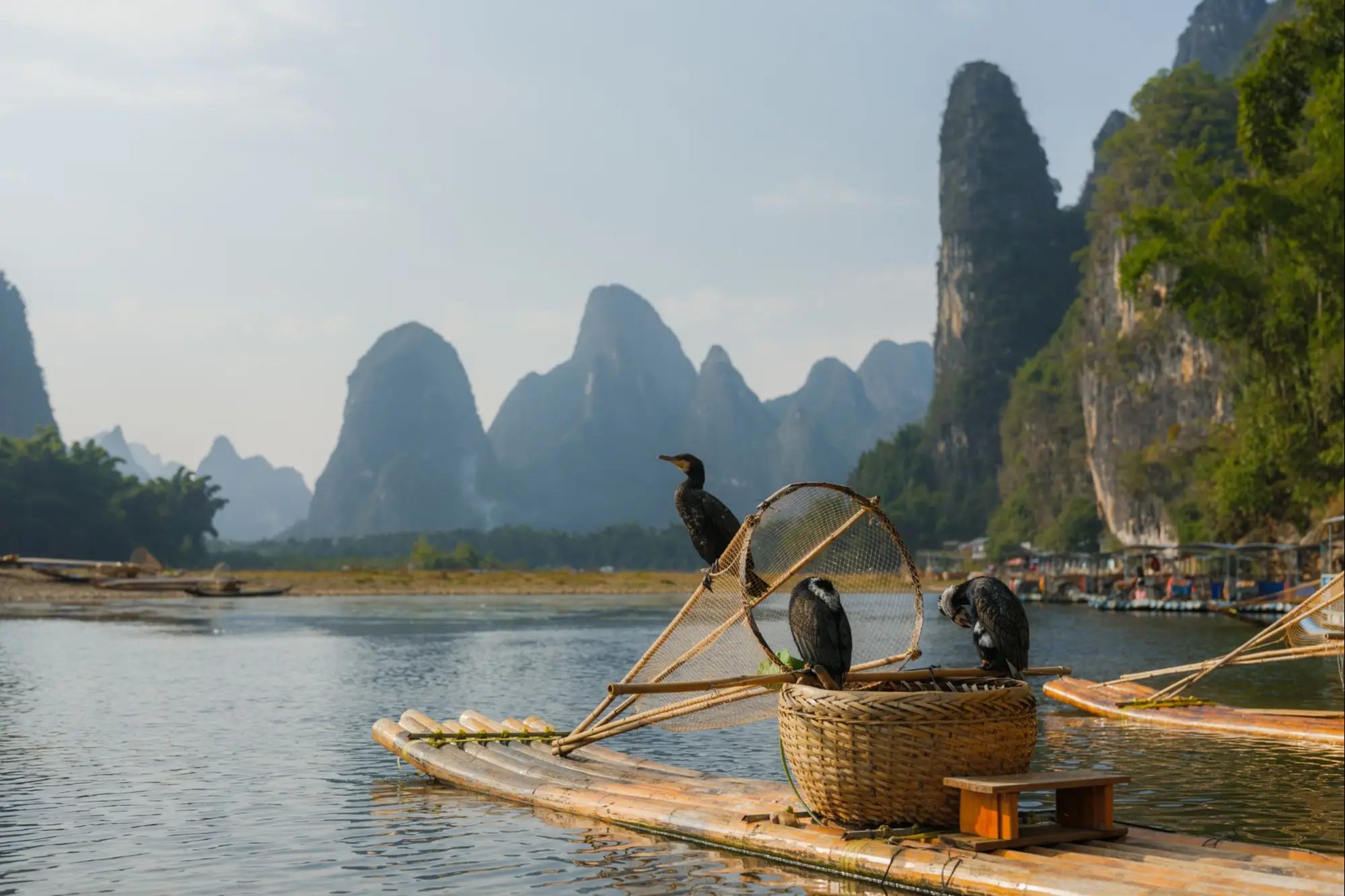 Kormoranfischer auf Bambusfloß am Li-Fluss in China mit dramatischen Karstbergen im Hintergrund bei Sonnenuntergang.