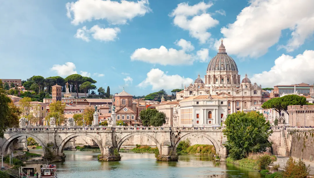 Panoramablick auf Rom mit Petersdom, historischer Brücke über den Tiber und blauem Himmel mit weißen Wolken.