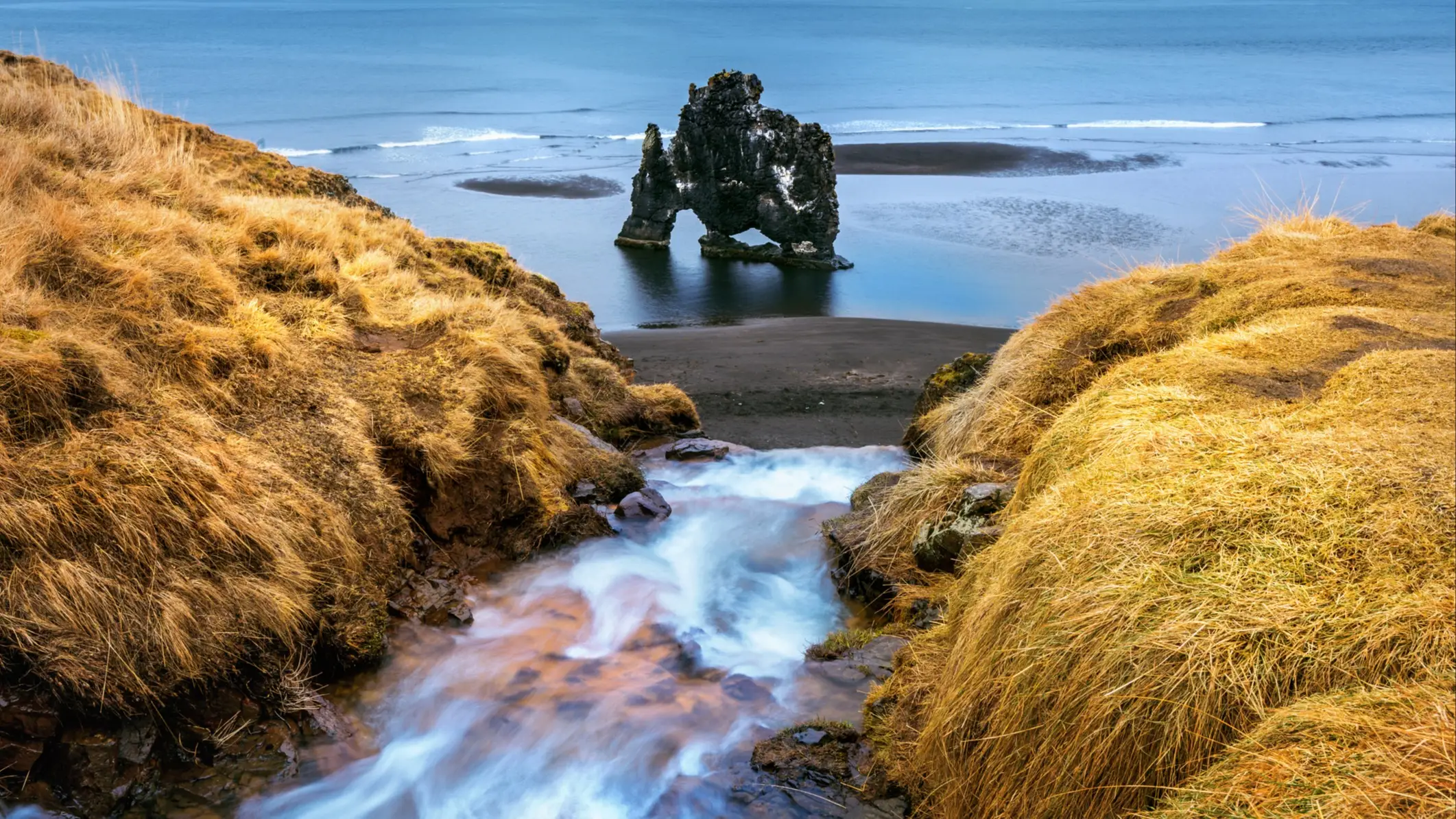 Formation rocheuse Hvítserkur dans l'océan en Islande, avec ruisseau cascadant entre collines d'herbe dorée.