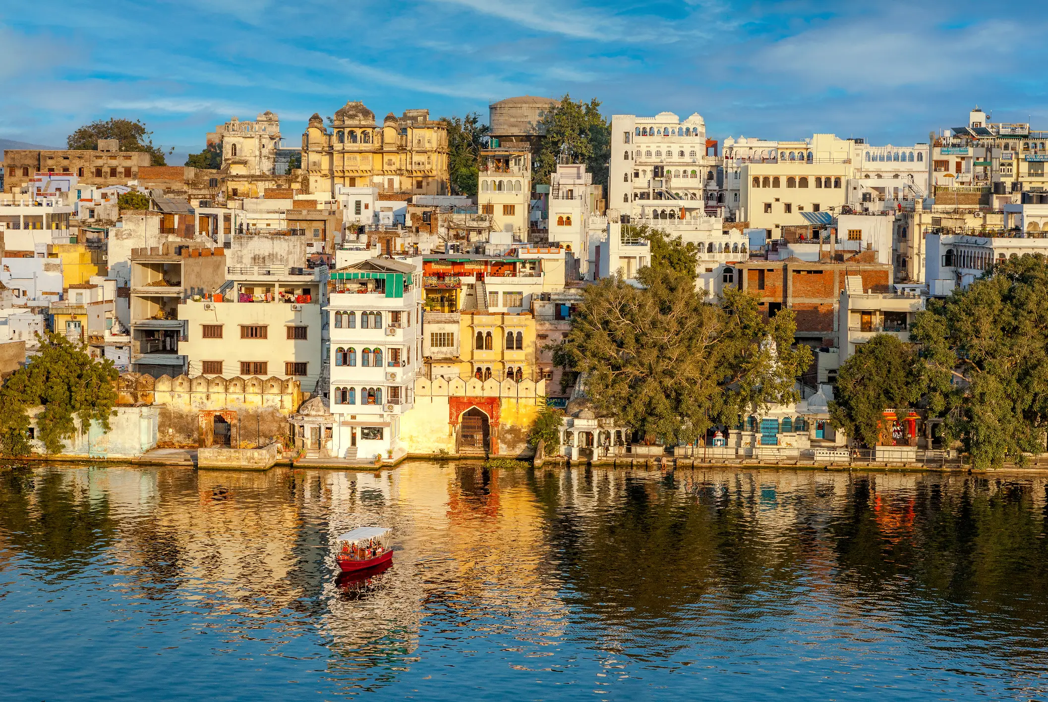 Historische Stadtansicht von Udaipur in Indien mit bunten Gebäuden am Wasser und einem kleinen roten Boot im Sonnenuntergangslicht.