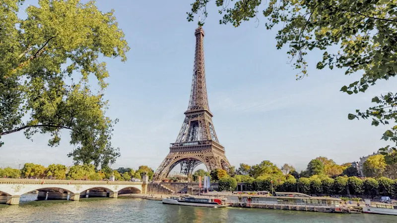 Eiffel Tower on a sunny day, framed by green trees, with Seine River and boats in the foreground under blue sky.