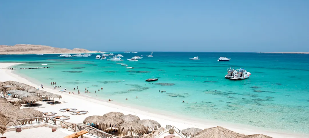 Tropischer Strand mit türkisblauem Wasser, weißem Sand, Strohschirmen und ankernden Booten unter klarem Himmel.