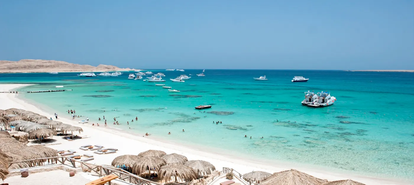 Tropischer Strand mit türkisblauem Wasser, weißem Sand, Strohschirmen und ankernden Booten unter klarem Himmel.