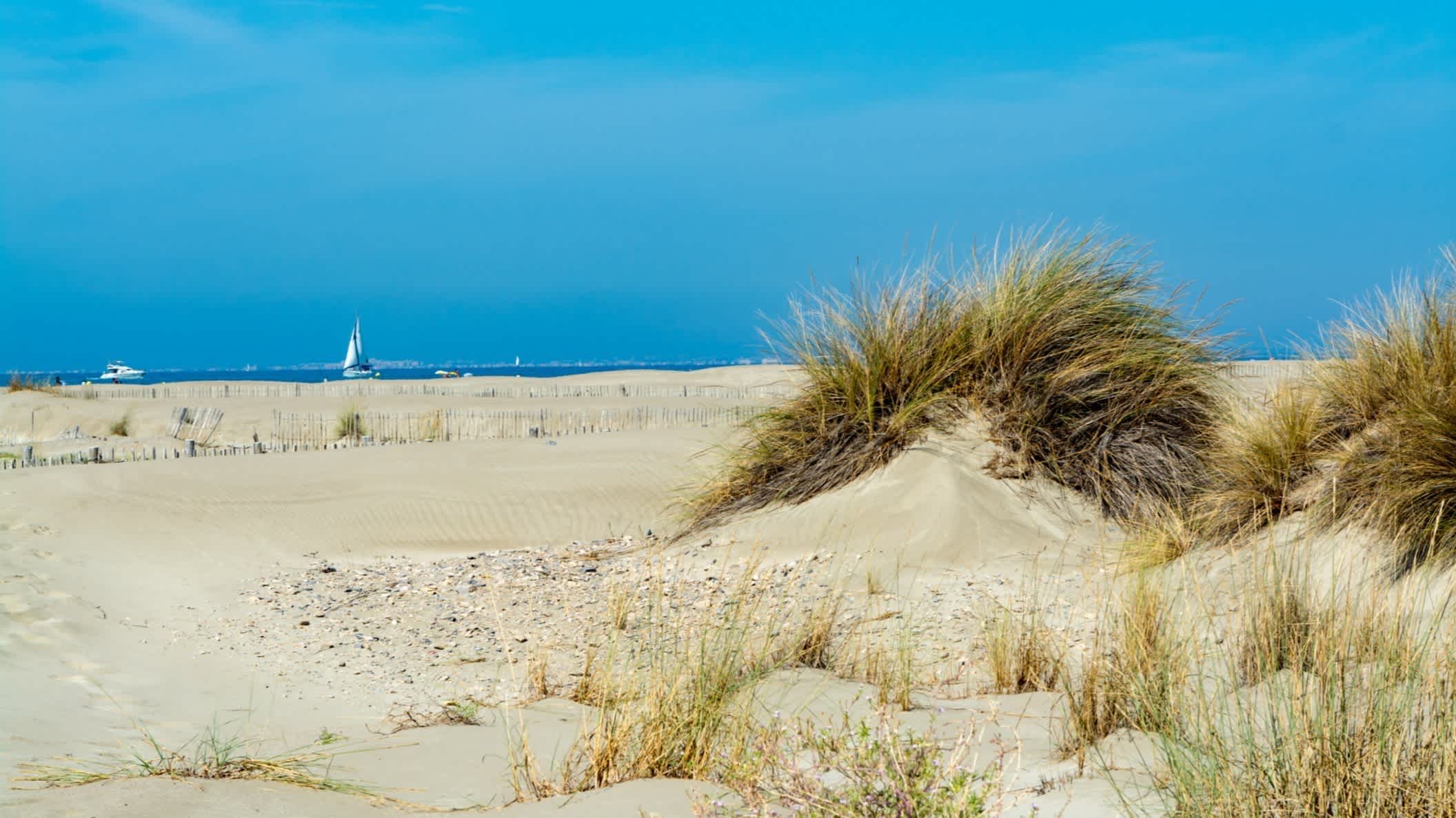 Der weiße Sandstrand Plage de Espiguette in Le Grau du Roi, Frankreich mit Aussicht auf die Dünen und das Meer am Horizont mit einem Segelboot.
