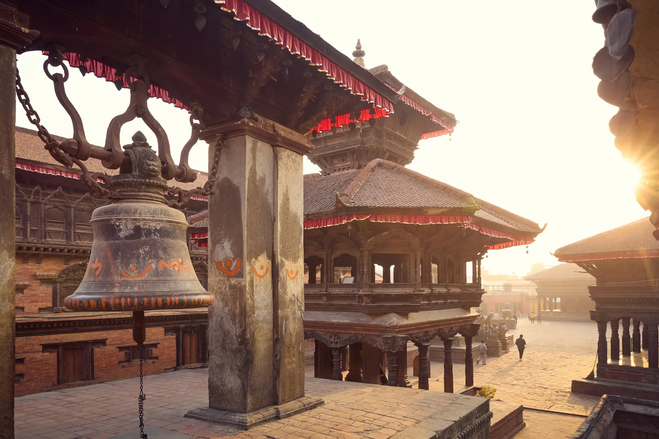 Historischer Tempel in Nepal mit großer Glocke im Vordergrund, traditionellen roten Dachkanten und goldener Abendsonne.