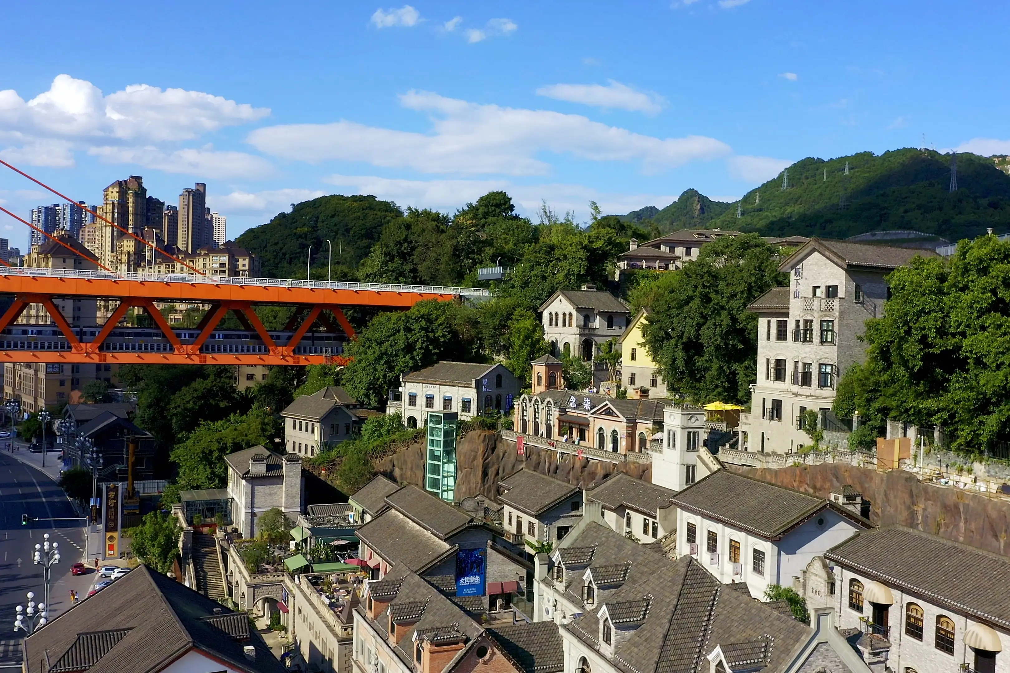 Eine leuchtend orangefarbene Brücke überspannt ein hügeliges Viertel mit grauen Gebäuden im traditionellen Stil, umgeben von üppigen grünen Bäumen und entfernten Hochhäusern unter einem klaren blauen Himmel.