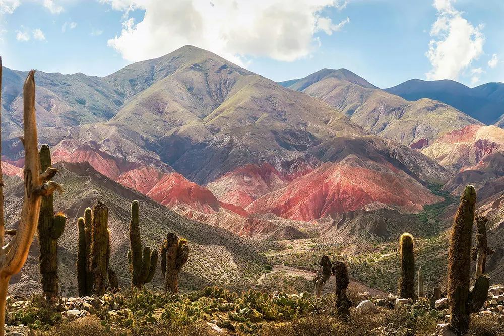 Chile Atacama desert diego-jimenez Eine Person steht allein in einer weiten, felsigen Landschaft mit rötlich-braunem Boden, weißem Sand und türkisfarbenem Wasser, umgeben von fernen Bergen unter einem strahlend blauen Himmel.