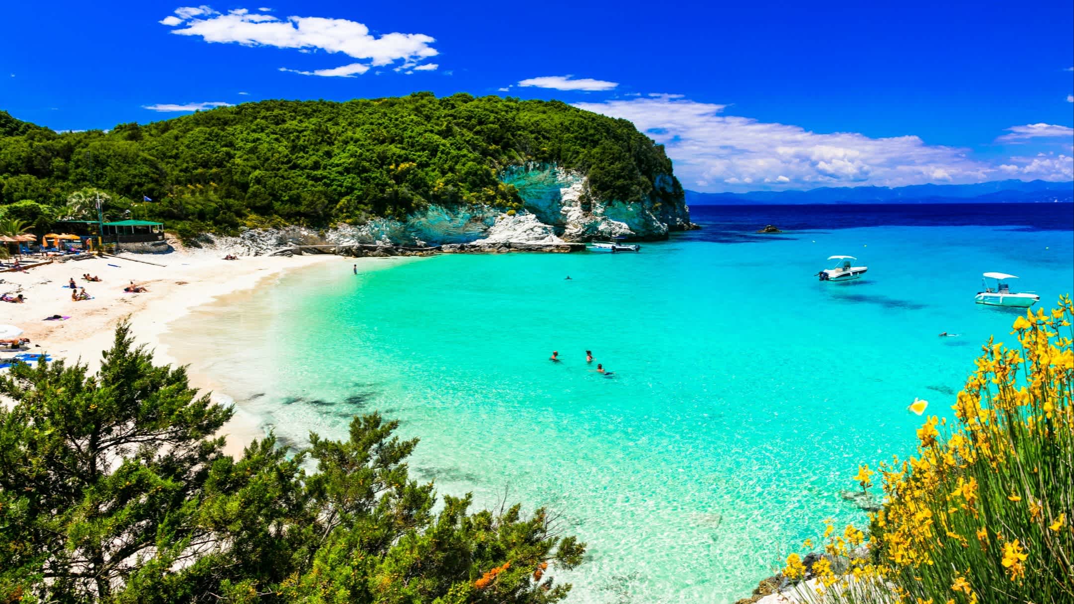 Der Strand von Vrika in Antipaxos, Ionische Inseln, Griechenland mit Blick über die gesamte Bucht, das türkisene Wasser sowie den belebten Strand und Booten.