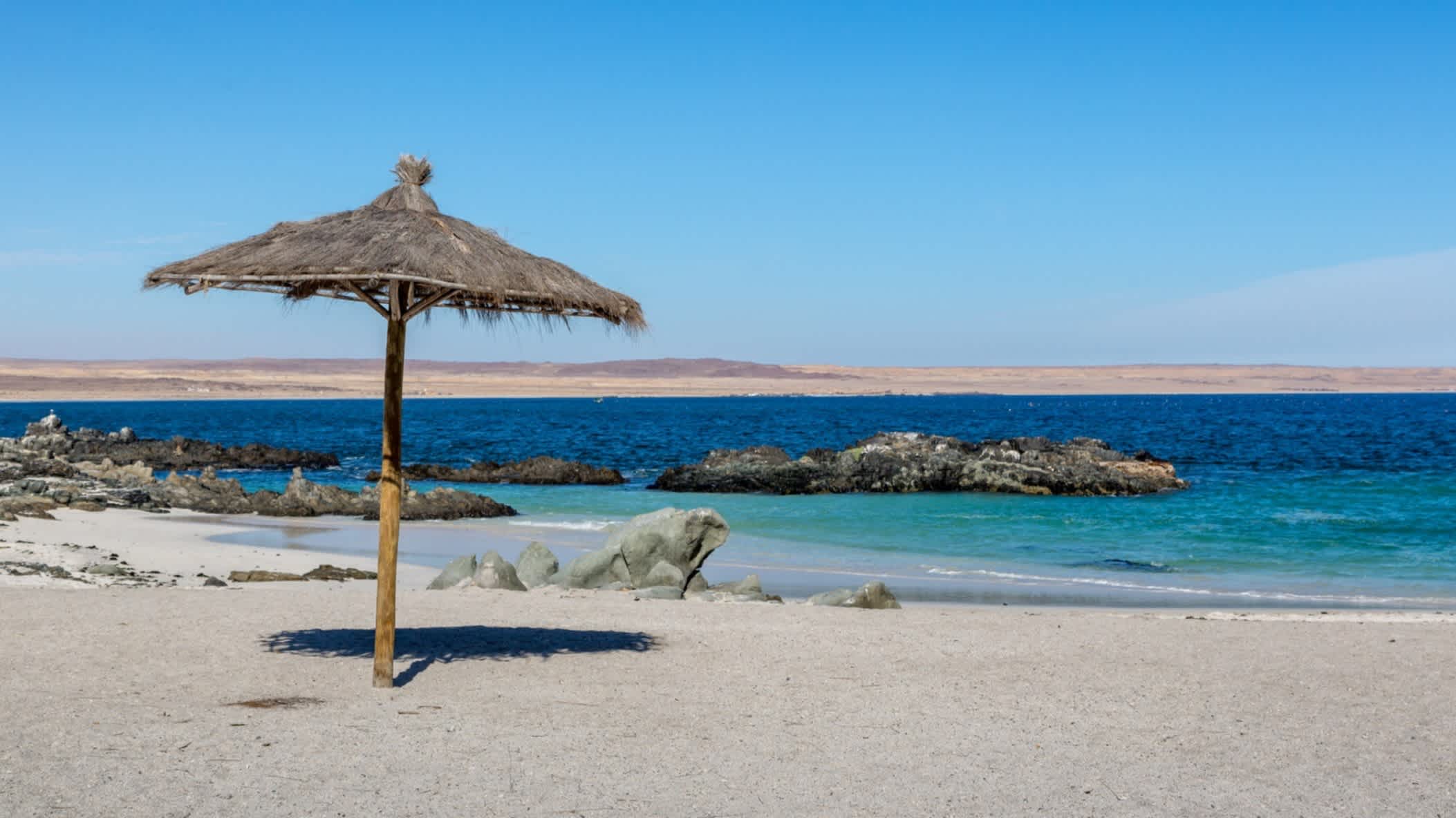 Ein Sonnenschirm steht am Strand der Bahia Inglesa in Chile nahe dem Ort Atacama und bei strahlendem Sonnenschein und azurblauem Wasser in einer sonst wüstenähnlichen Umgebung. 