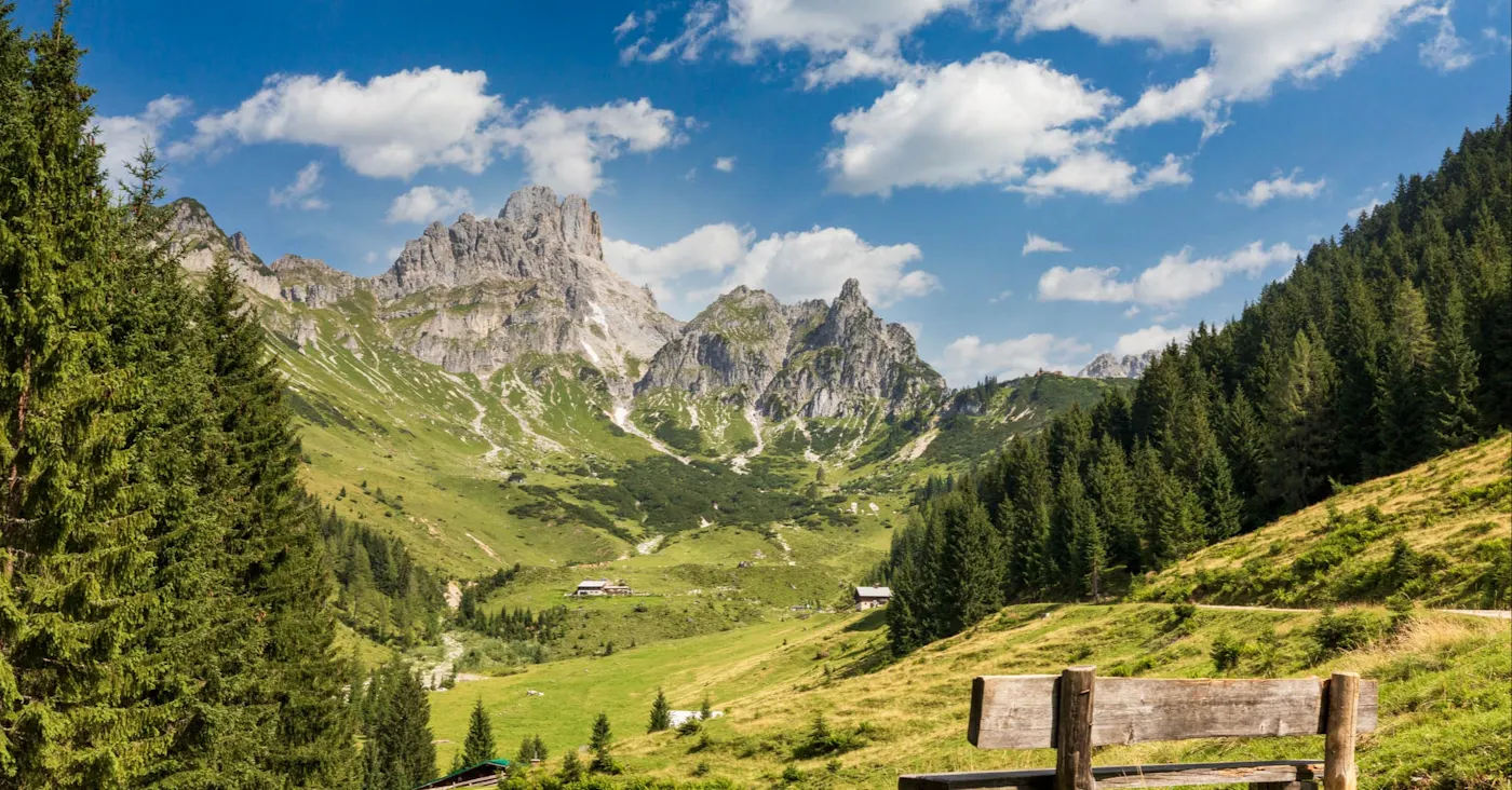 Wooden bench overlooking Alpine valley with jagged mountain peaks, green meadows, and evergreen forests under blue sky.
