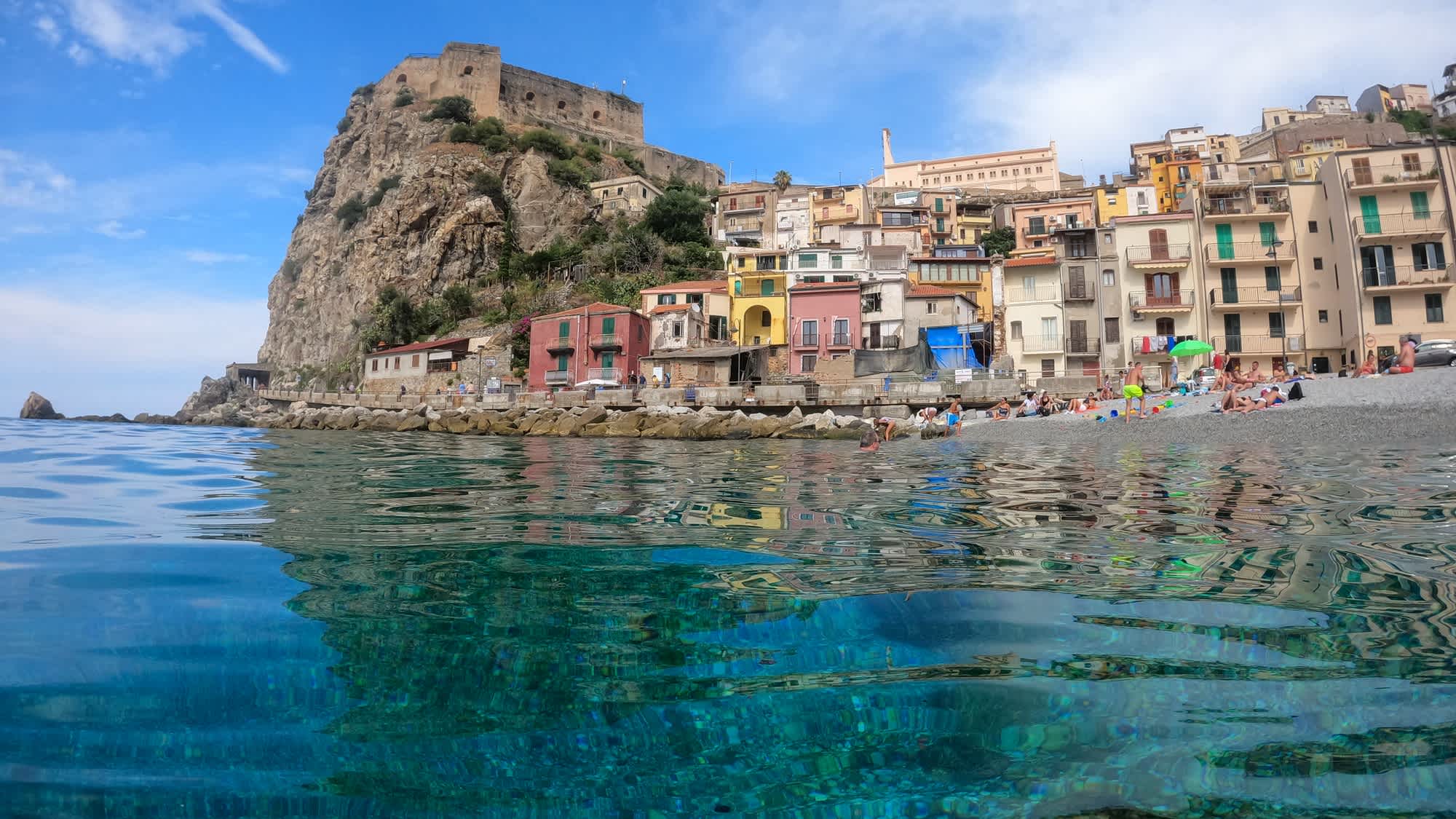 Blick auf den Strand von Scilla, Kalabrien, mit bunten Häusern und einer Felsenfestung im Hintergrund.