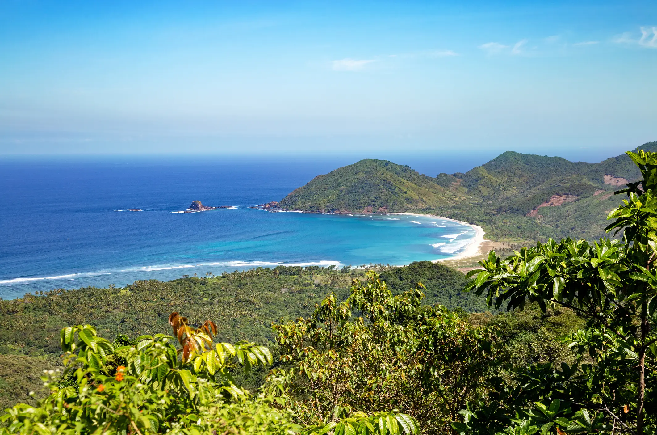 Panoramablick auf eine tropische Küste mit türkisblauem Wasser, weißem Sandstrand und grünen Hügeln unter klarem Himmel.
