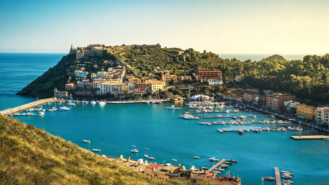 Hafen mit bunten Häusern und grüner Halbinsel. Monte Argentario, Toskana, Italien.