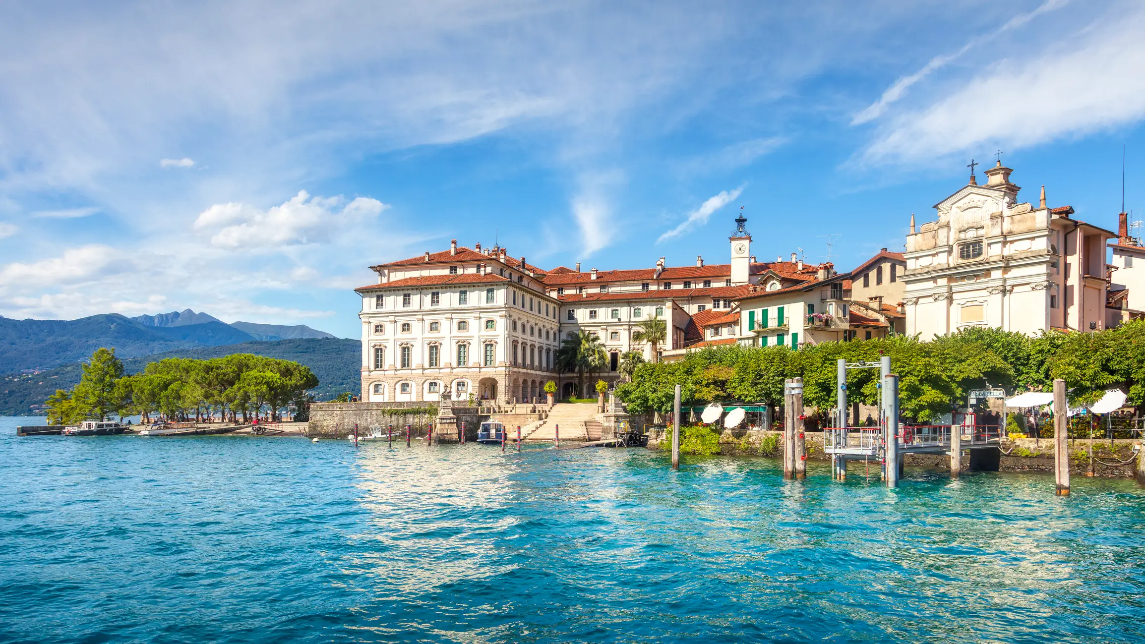 Verbania Elegante historische Gebäude und eine Kirche liegen am klaren blauen Wasser, umgeben von üppigem Grün und fernen Bergen unter einem sonnigen Himmel am Lago Maggiore in Italien.