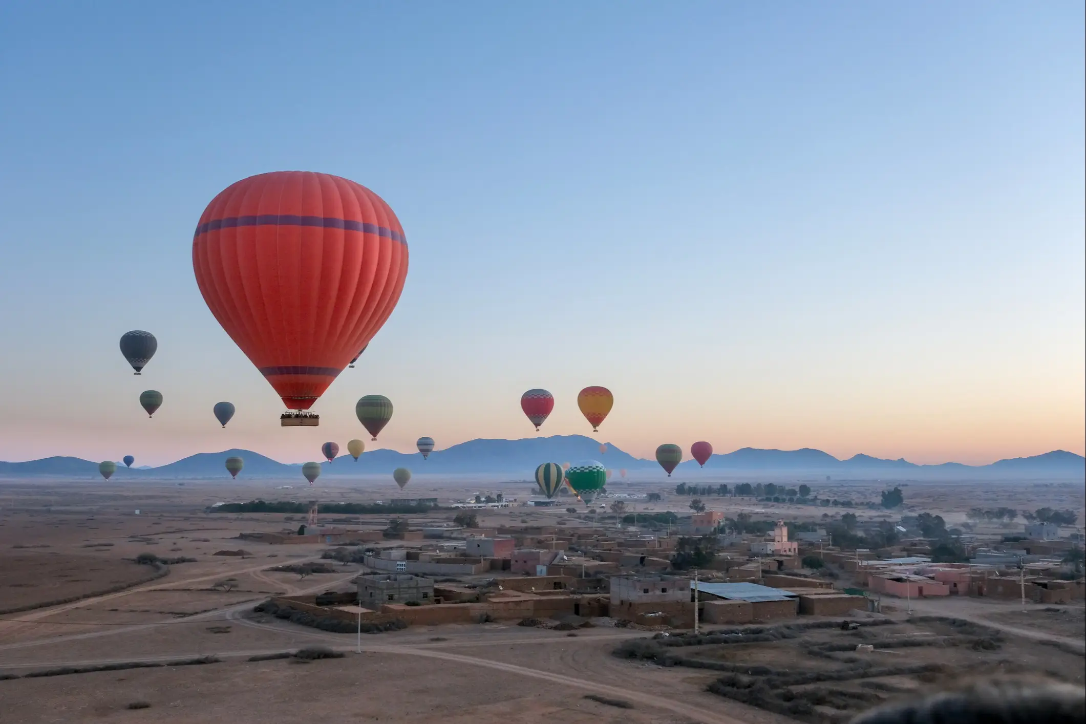 Bunte Heißluftballons schweben über einer Wüstenlandschaft mit kleinem Dorf bei Sonnenaufgang, großer roter Ballon im Vordergrund.