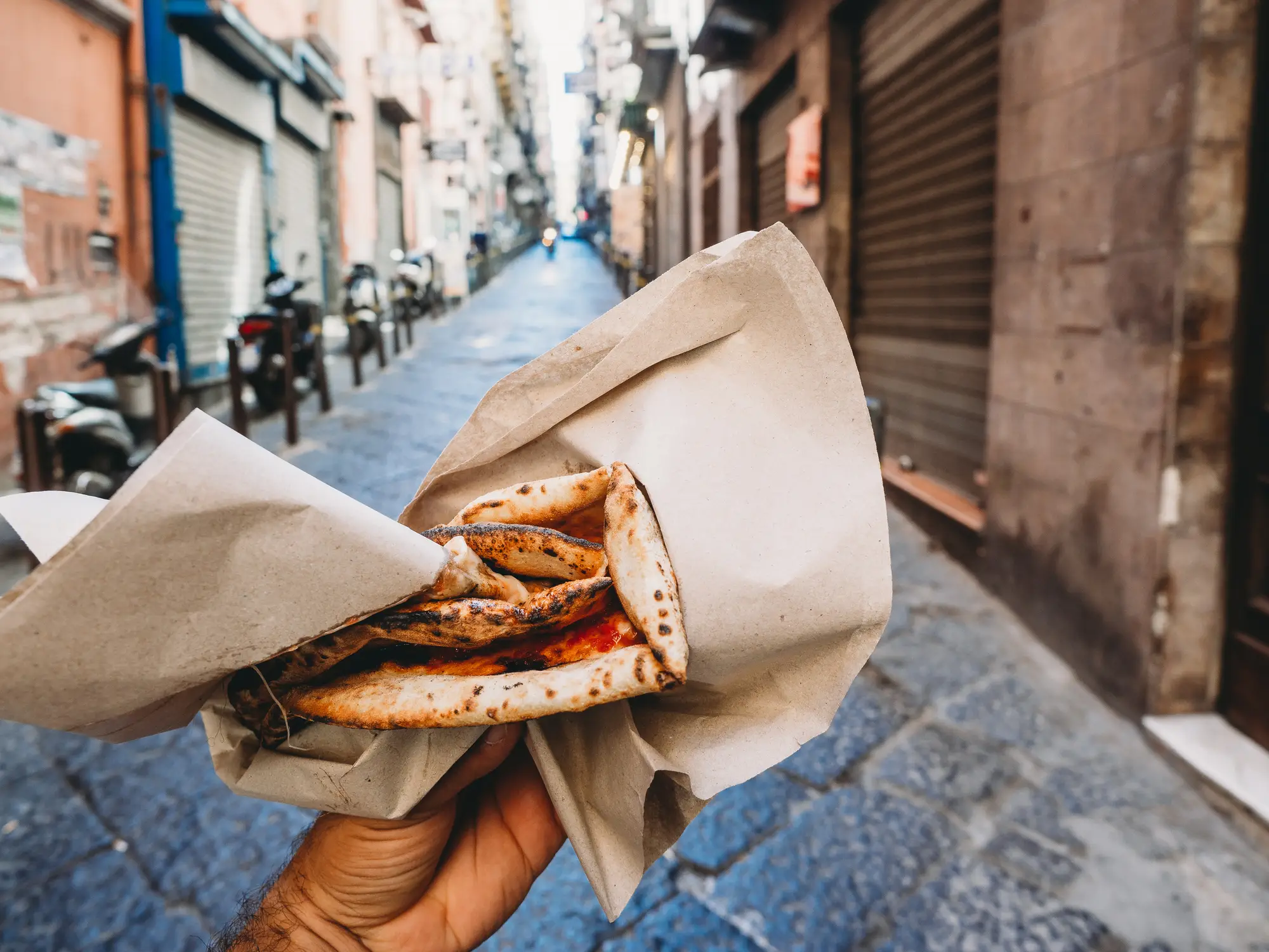 Eine Hand hält eine gefaltete, in braunes Papier eingewickelte Pizza in einer schmalen, leeren Straße, die von verschlossenen Geschäften und Motorrollern gesäumt ist. Die Szene erinnert an ein zwangloses Essen in einer europäischen Stadt.