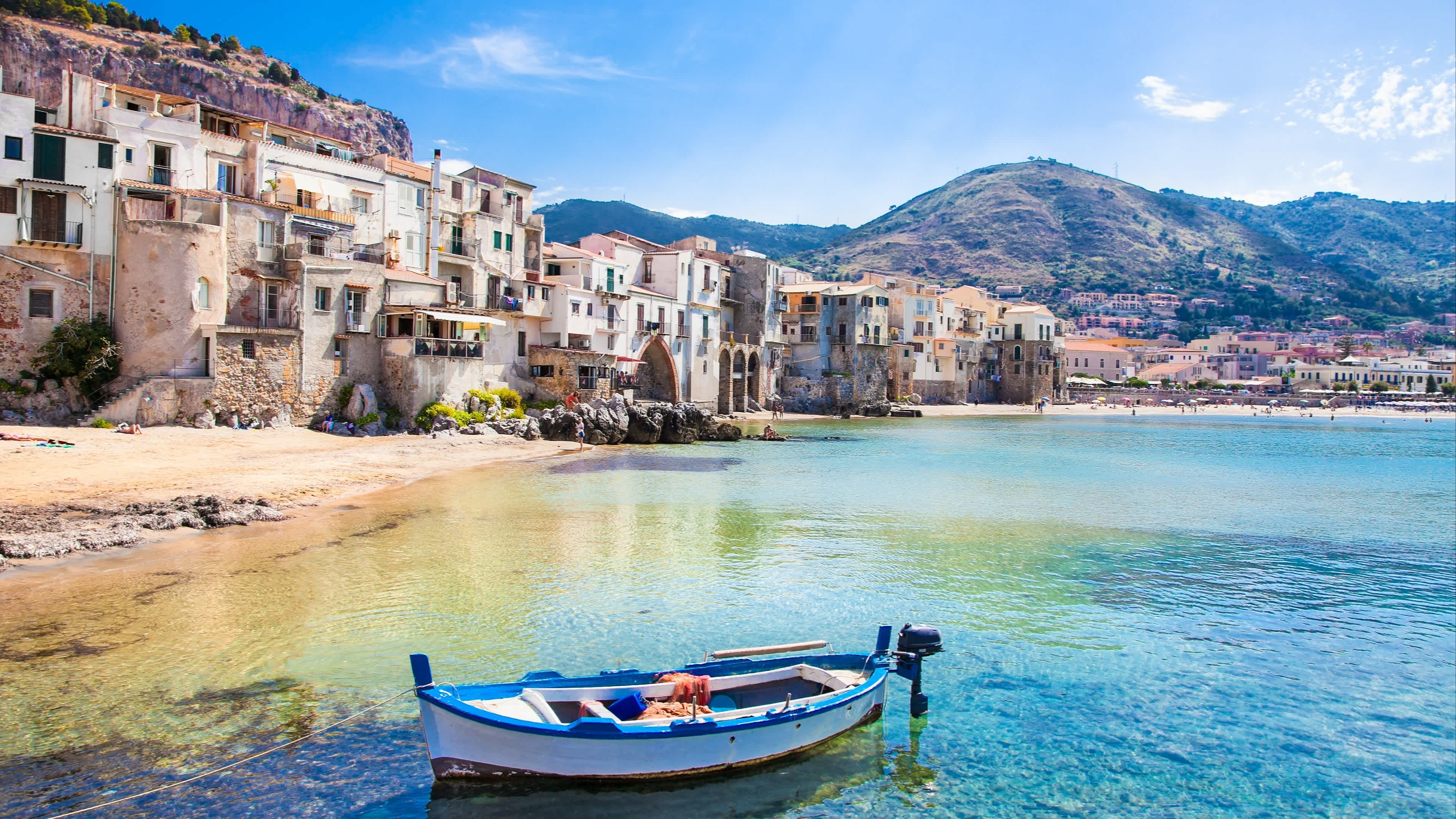 Schöner alter Hafen mit hölzernem Fischerboot in Cefalu, Sizilien, Italien mit Häusern, Klippen und Bergen im Bild bei Sonnenschein.

