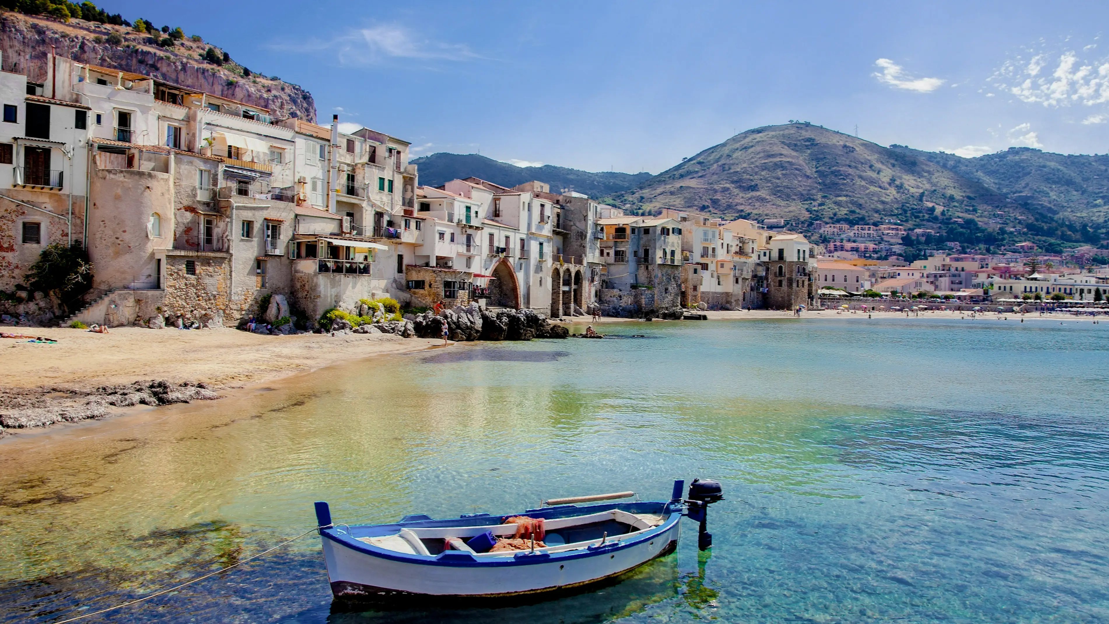 Schöner alter Hafen mit hölzernem Fischerboot in Cefalu, Sizilien, Italien mit Häusern, Klippen und Bergen im Bild bei Sonnenschein.

