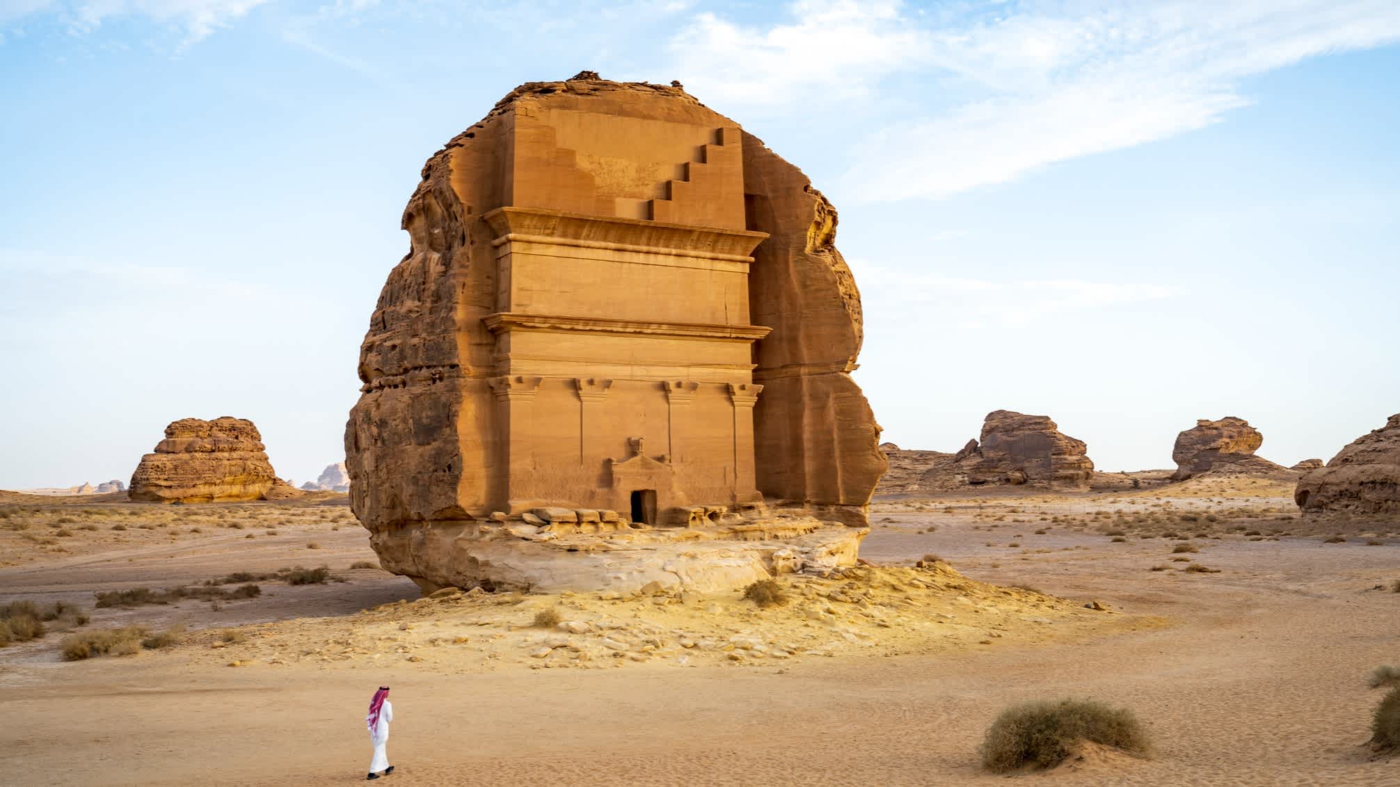 Ein Mann mit langer weißer Robe vor einem großen felsengrab in der Wüste