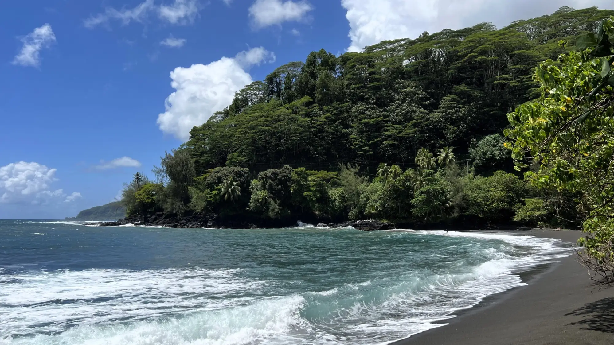 Plage de sable noir avec vagues turquoise bordée d'une forêt tropicale luxuriante sous un ciel bleu parsemé de nuages blancs.