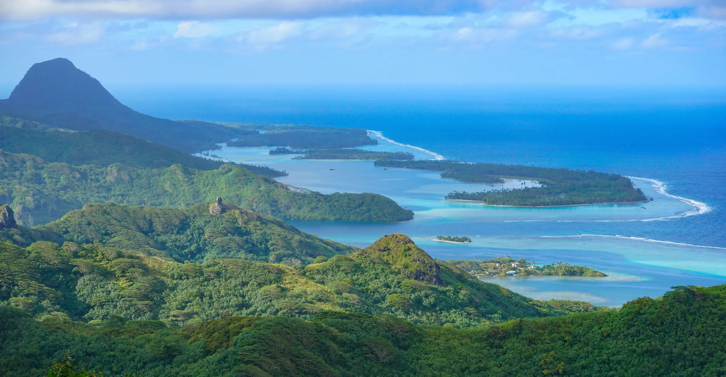 Huahine, Polynésie Française Des collines verdoyantes et luxuriantes descendent vers un littoral turquoise parsemé de lagunes sinueuses et de récifs coralliens, bordé par l'océan bleu sous un ciel partiellement nuageux, avec une montagne en arrière-plan.