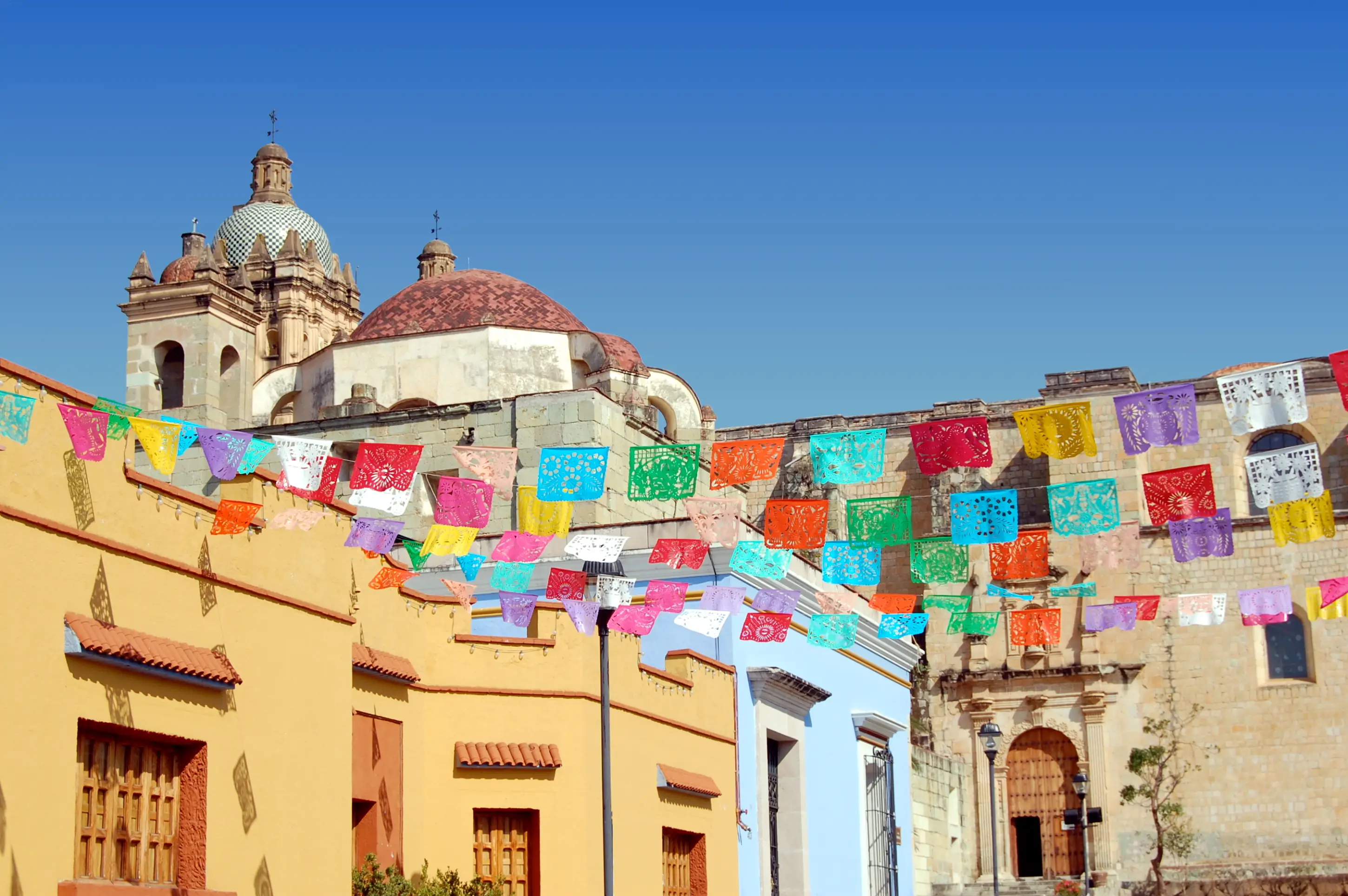 Historische mexikanische Kirche mit Kuppeln unter blauem Himmel, bunte Papierwimpel schmücken die Straße vor gelben Gebäuden.