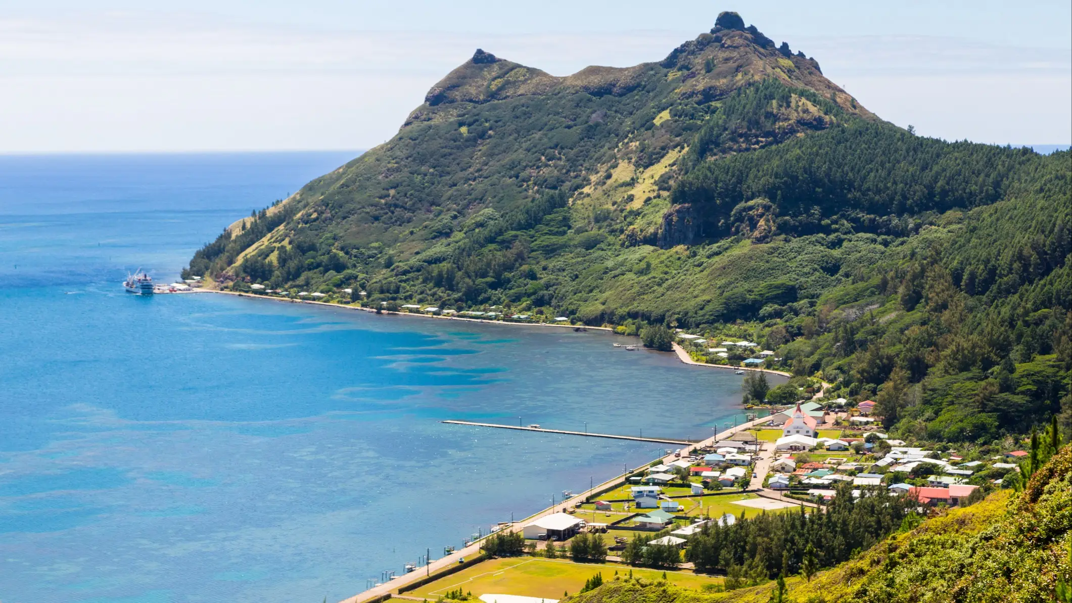 Küstenlandschaft mit grünem Berg, türkisblauem Meer und kleiner Siedlung am Ufer unter klarem Himmel.