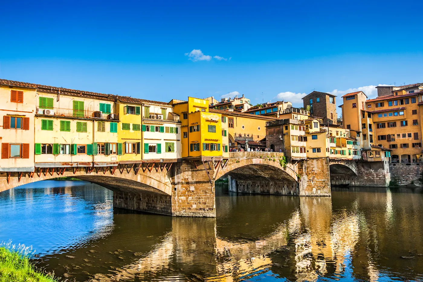Ponte Vecchio Bridge, Florence Farbenfrohe Gebäude säumen die historische Brücke Ponte Vecchio über den Arno in Florenz, Italien, unter einem strahlend blauen Himmel, der sich im ruhigen Wasser spiegelt.