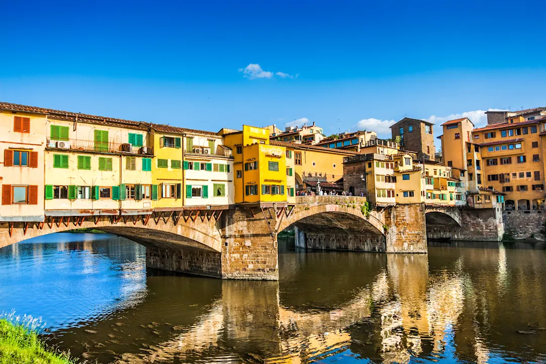 Farbenfrohe Gebäude säumen die historische Brücke Ponte Vecchio über den Arno in Florenz, Italien, unter einem strahlend blauen Himmel, der sich im ruhigen Wasser spiegelt.