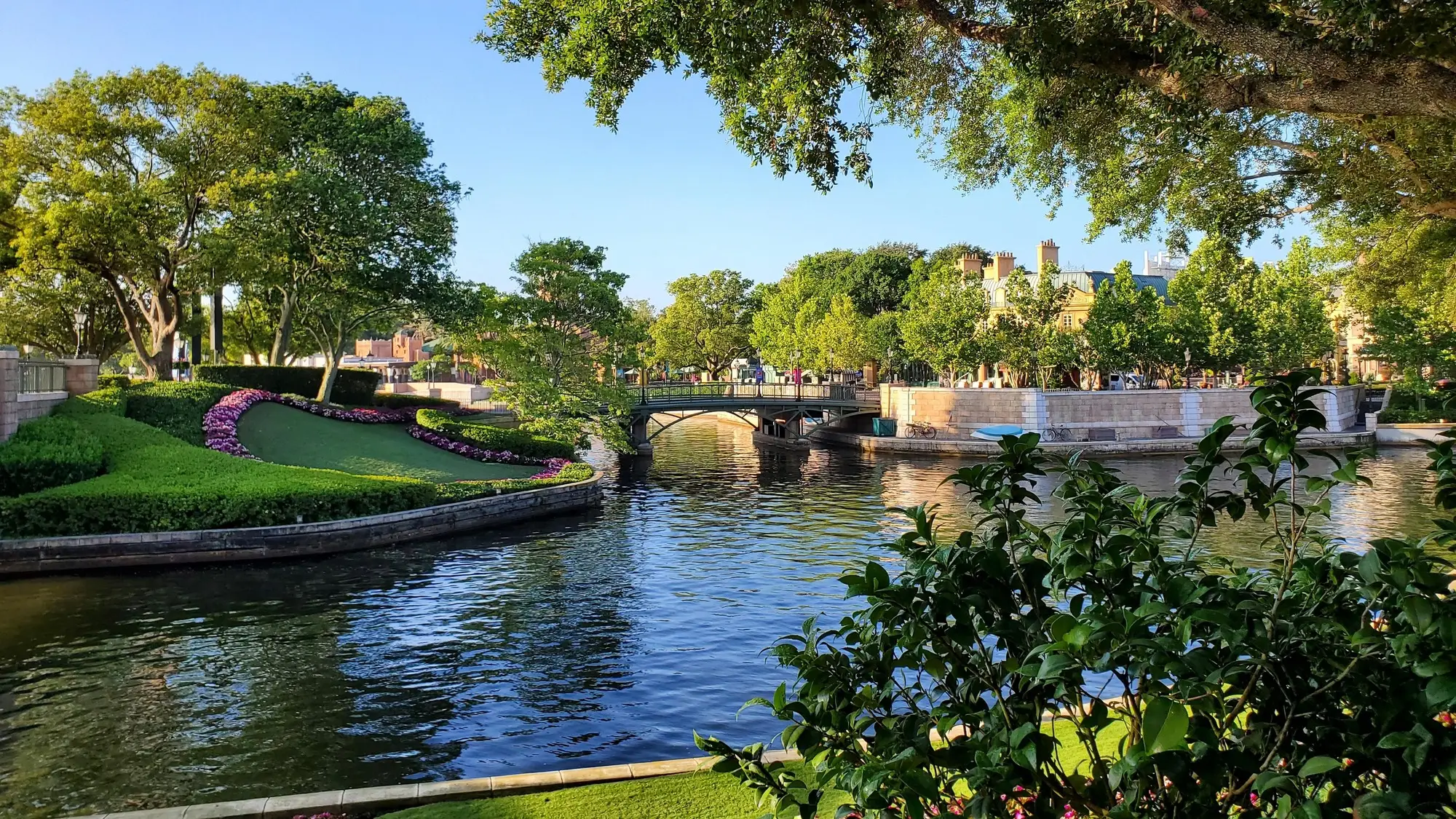 Parc pittoresque avec canal d'eau, pont élégant, jardins fleuris et arbres verdoyants sous un ciel bleu ensoleillé.