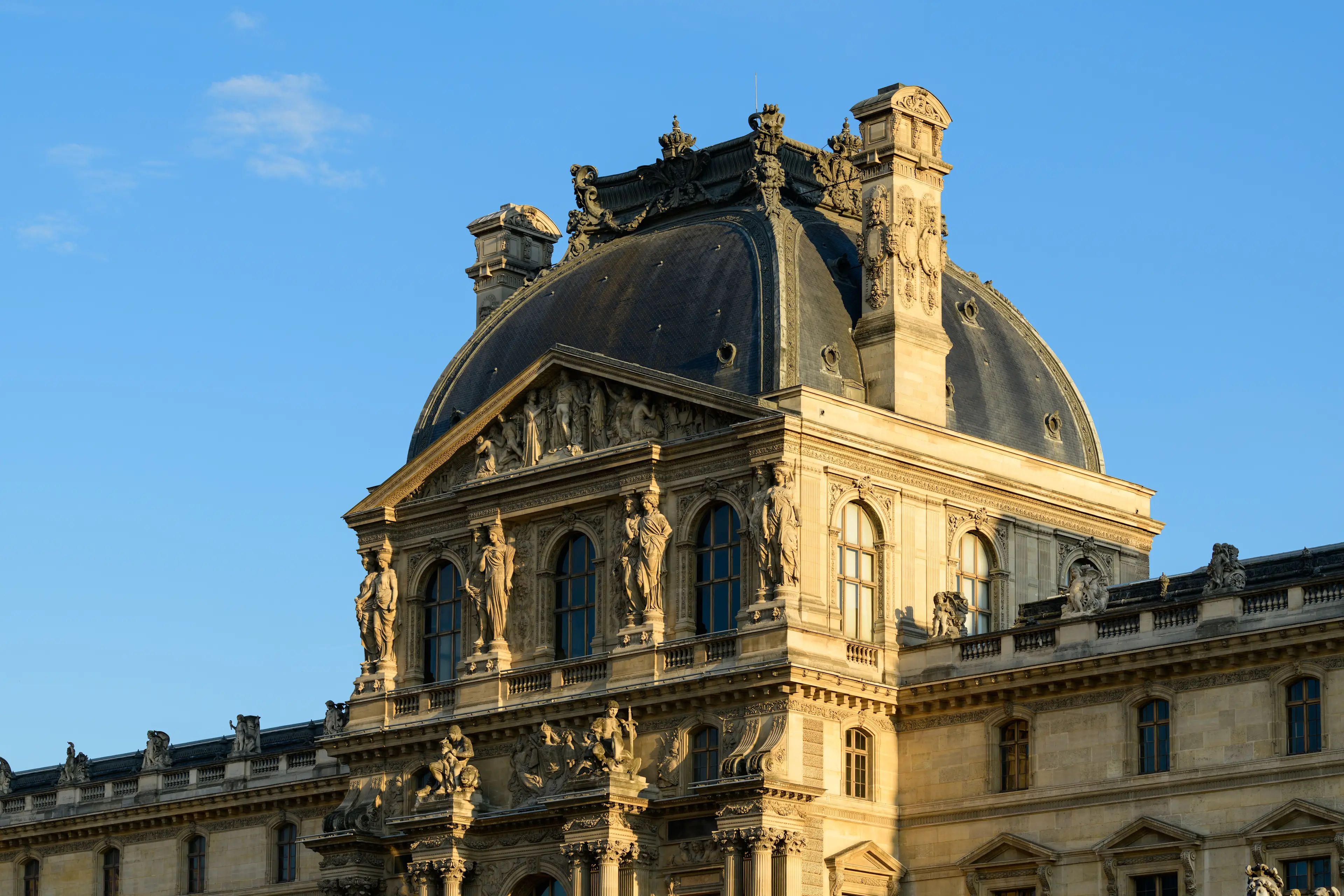 Ornate section of the Louvre Museum with its distinctive dark domed roof and classical statues against a clear blue sky.