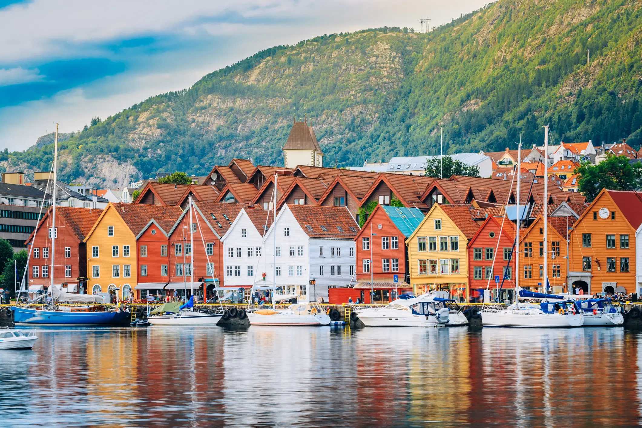 Bunte Holzhäuser säumen die Uferpromenade vor Segelbooten, mit grünen Hügeln und einem bewölkten Himmel im Hintergrund, in Bryggen, Bergen, Norwegen.