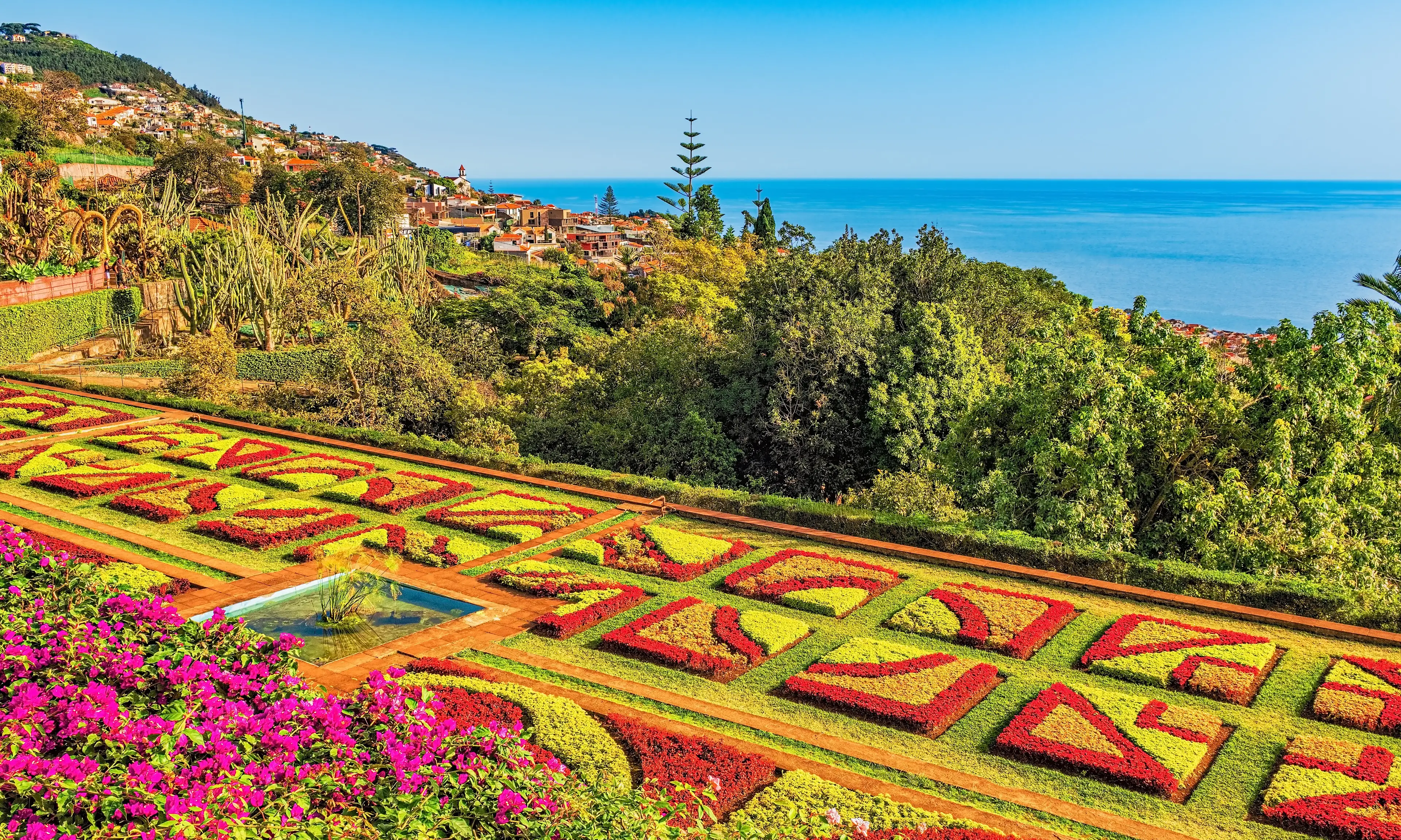 Botanischer Garten in Funchal, Madeira Bunte geometrische Blumenbeete in einem botanischen Garten mit Blick auf üppiges Grün und eine Küstenstadt, mit dem blauen Meer und dem klaren Himmel im Hintergrund. Zwischen den lebhaft gemusterten Beeten befindet sich ein kleines Wasserspiel.